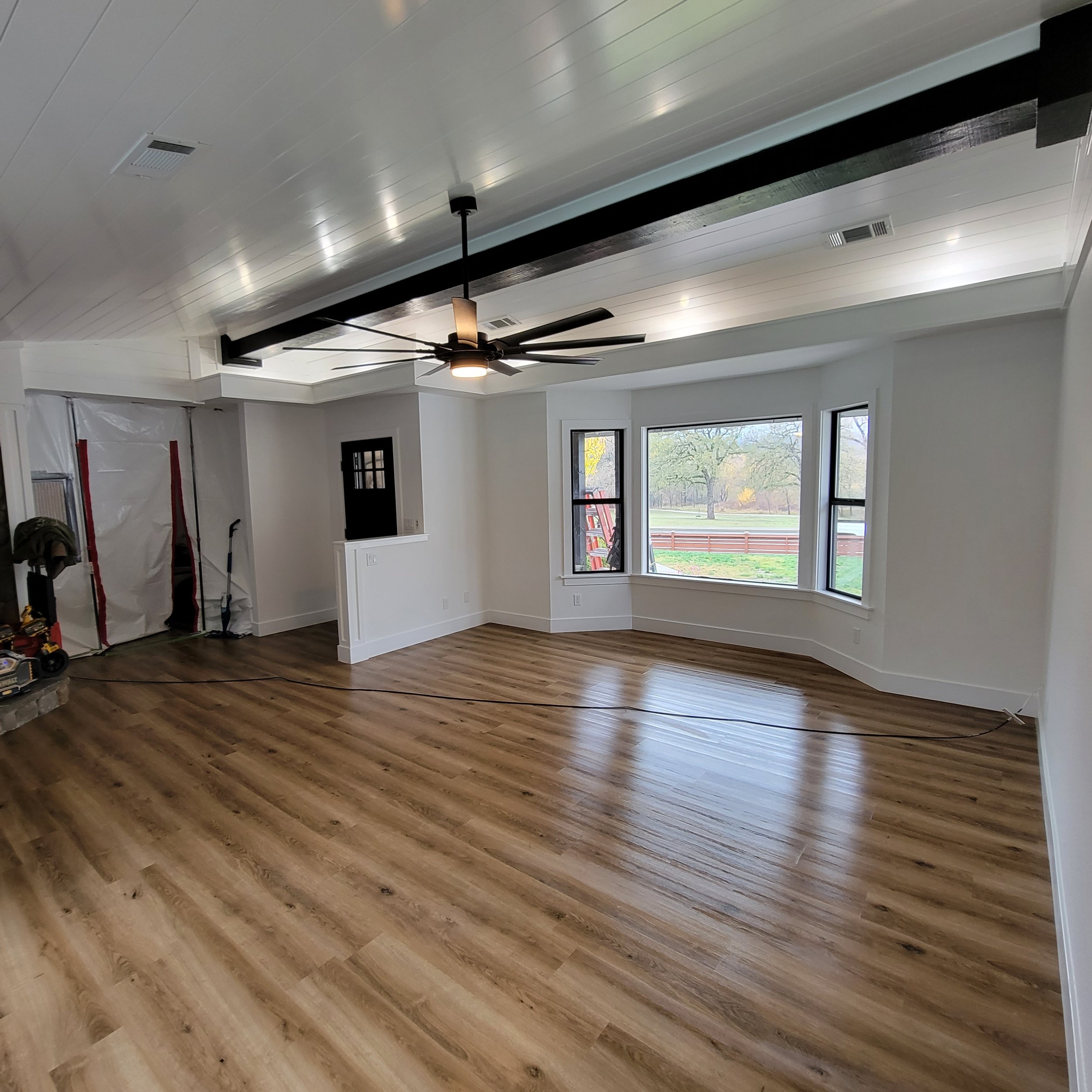 Empty living room with hardwood floors, white walls, large bay windows, a black ceiling fan, and a small black door. Construction tools and materials are visible in the corner.