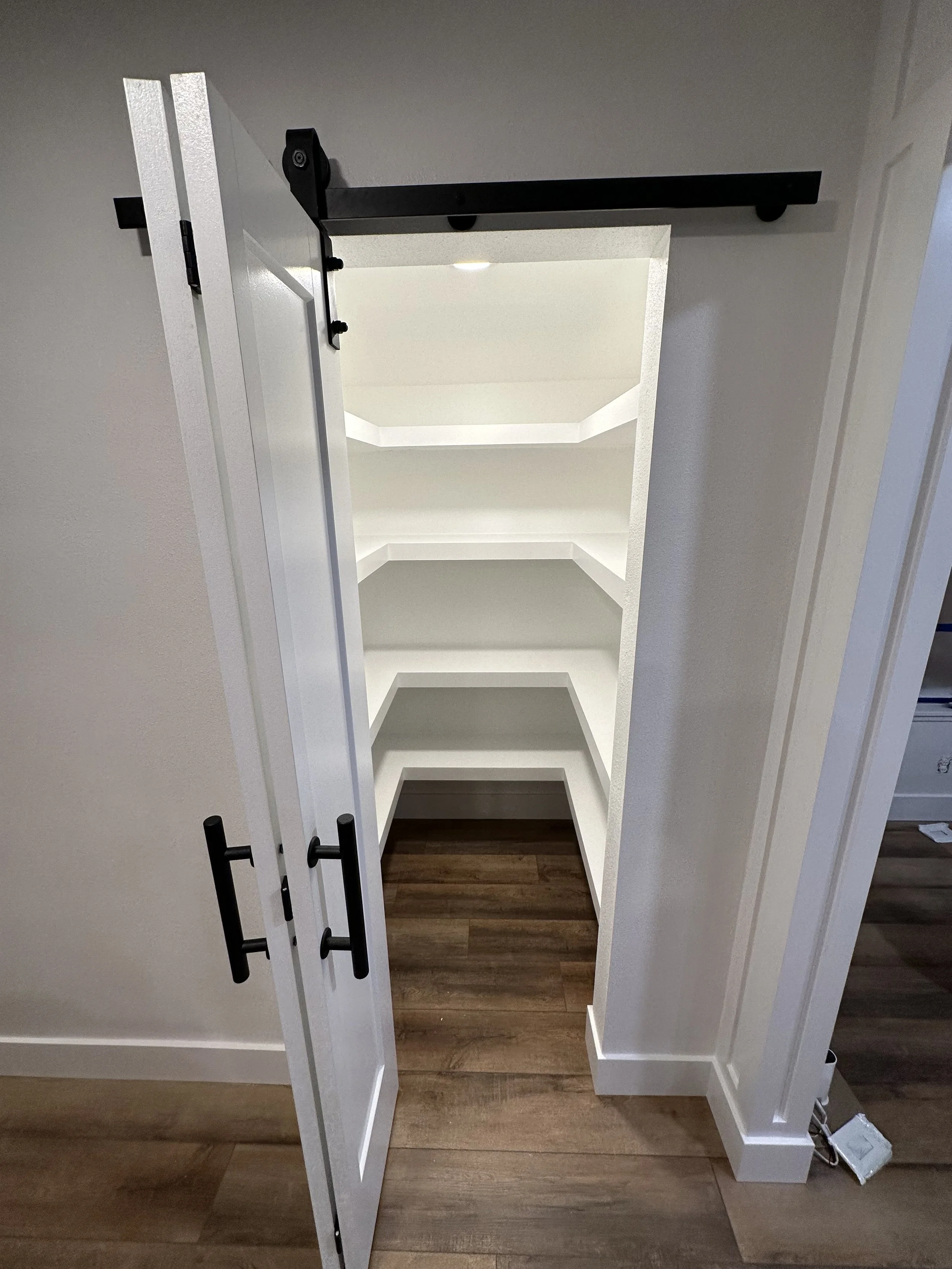 Inside a small pantry with white shelves, black sliding barn door hardware at the top, and a sliding white door with black handles, wood flooring, and a light grey wall.