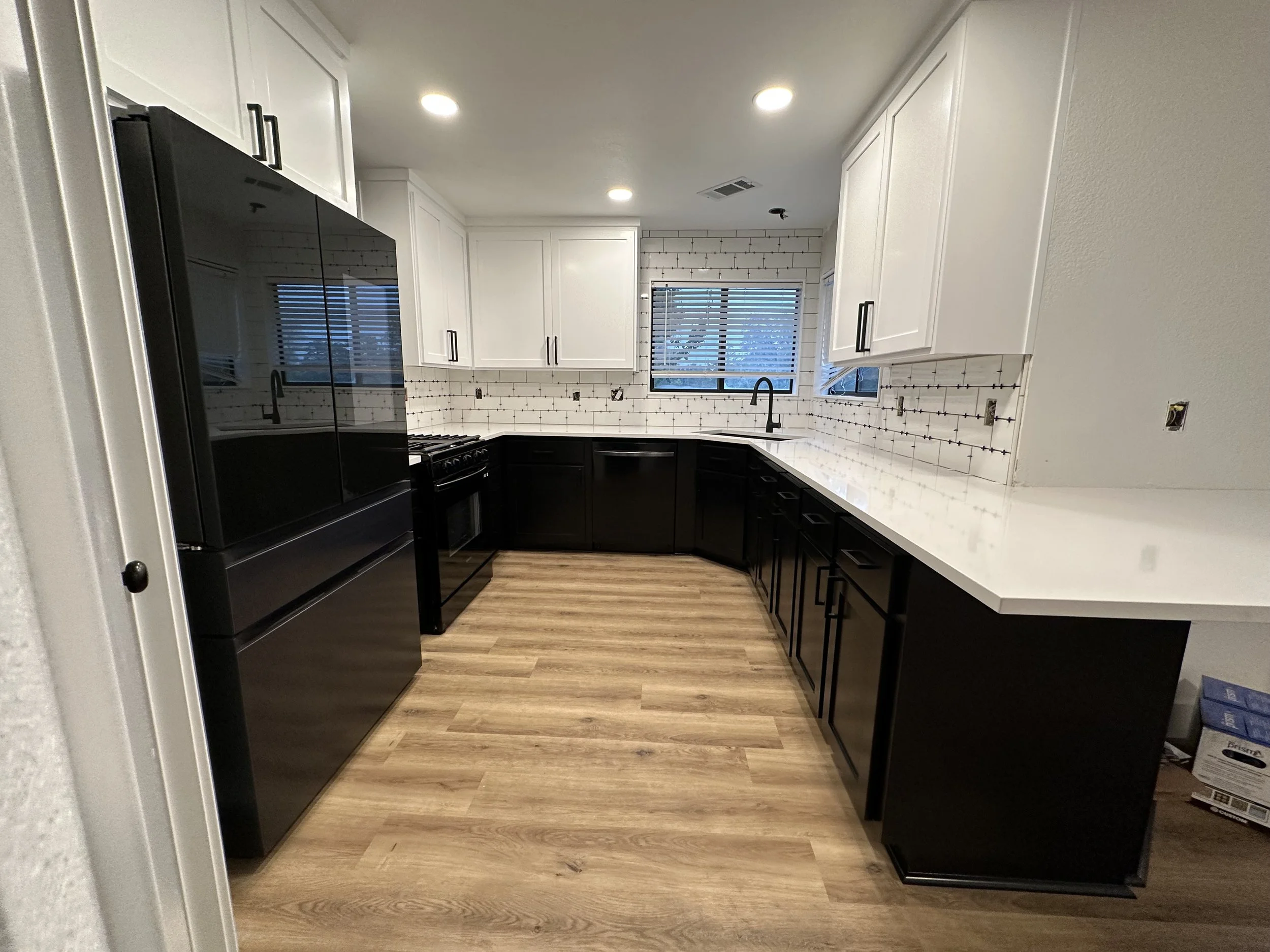 Modern kitchen with black lower cabinets, white upper cabinets, wooden flooring, and a white countertop. There is a black refrigerator, stove, and a black faucet at the sink, with a window and a subway tile backsplash.