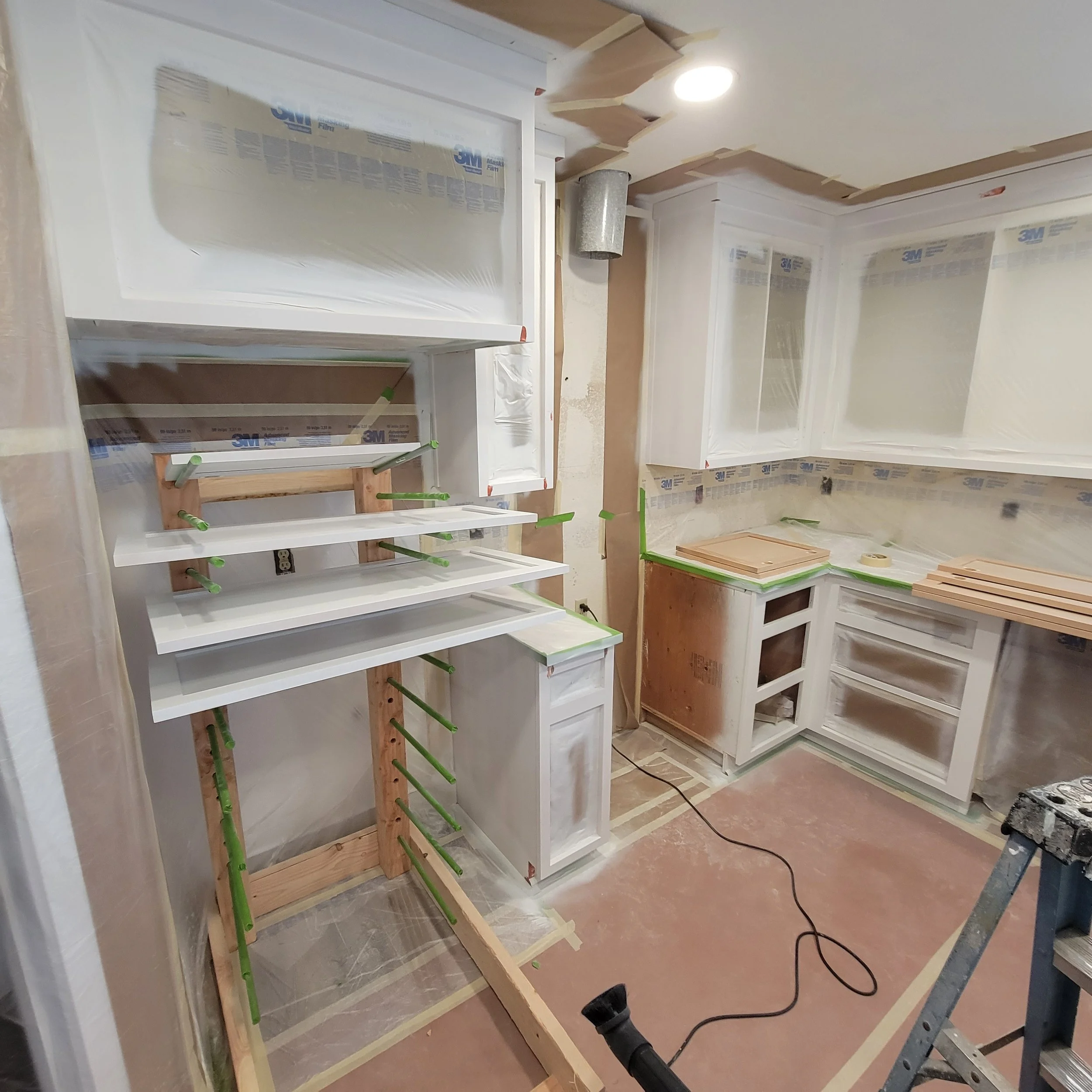 Kitchen under renovation with white cabinets, some with glass doors, partially painted walls, construction tape, ladder, and tools