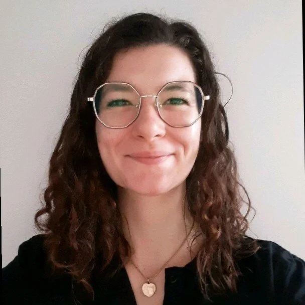 Jeune femme souriante avec des lunettes rondes, cheveux bruns bouclés, portant un collier en forme de cœur, contre un fond blanc.