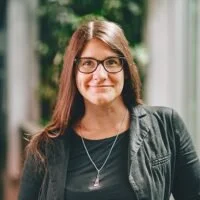 Femme souriante avec des lunettes, portant un vêtement noir, devant un mur de végétation à l'intérieur.
