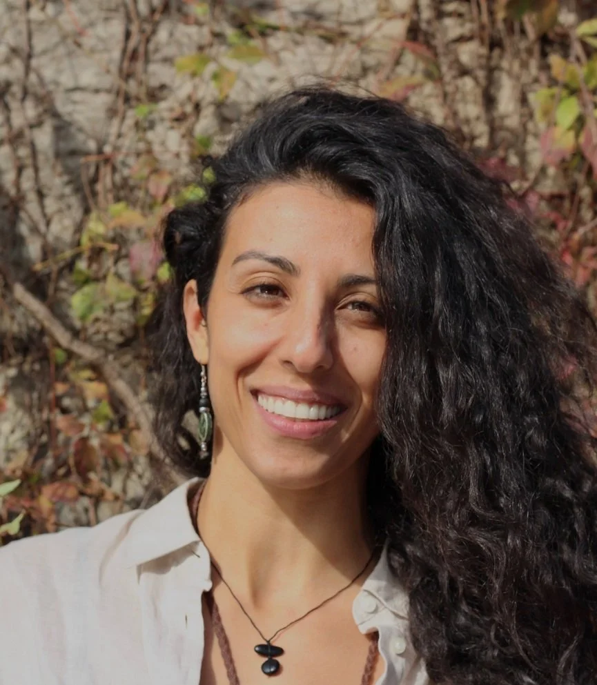 Une femme souriante avec des cheveux bouclés noirs, portant des boucles d'oreilles, un collier noir, et une chemise blanche, posant devant des arbustres.