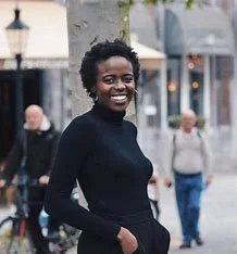 Femme souriante avec cheveux bouclés, portant un pull noir, dans une rue urbaine