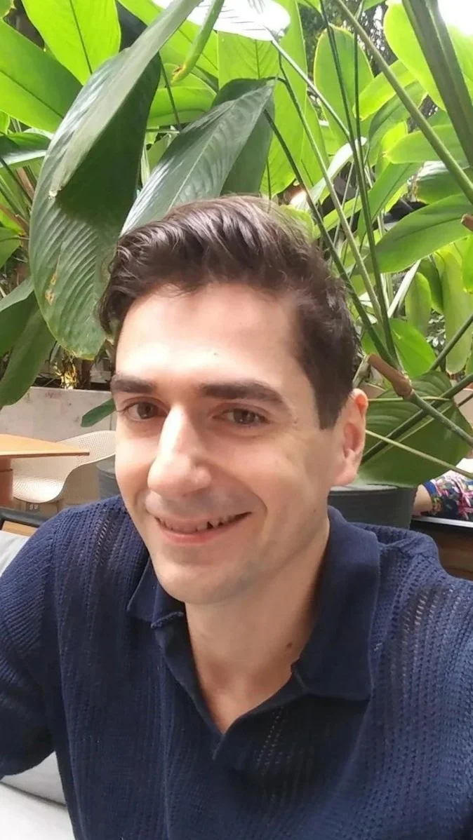 Un homme souriant, assis dans un environnement tropical avec beaucoup de grandes feuilles vertes en arrière-plan.