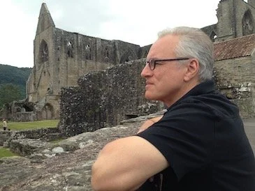 Homme portant des lunettes et un t-shirt noir, regardant vers la gauche, avec une vieille église en ruines en arrière-plan.