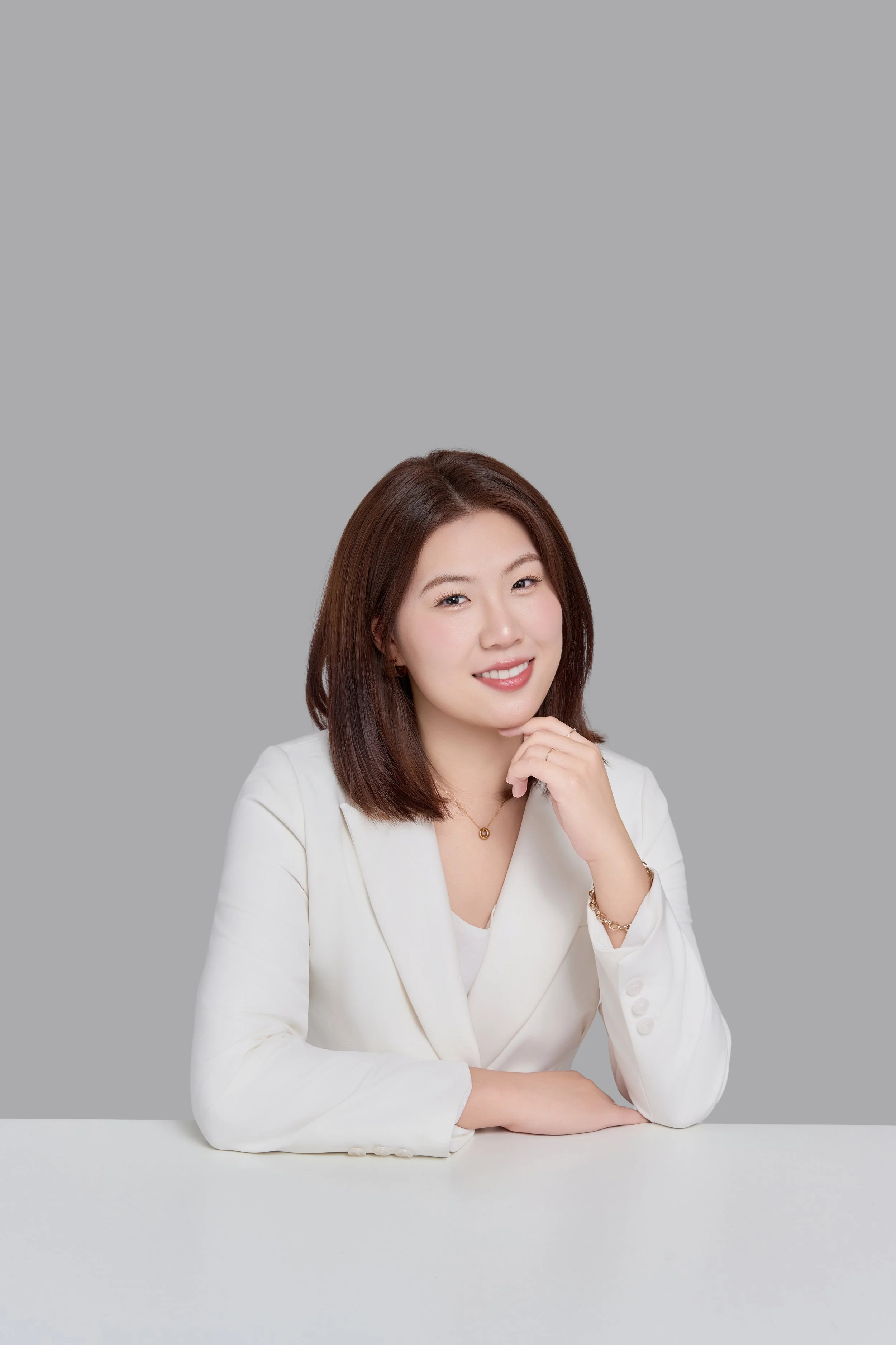 Une femme souriante avec des cheveux bruns, vêtue d'une veste blanche, posant avec sa main sous son menton contre un fond gris clair.