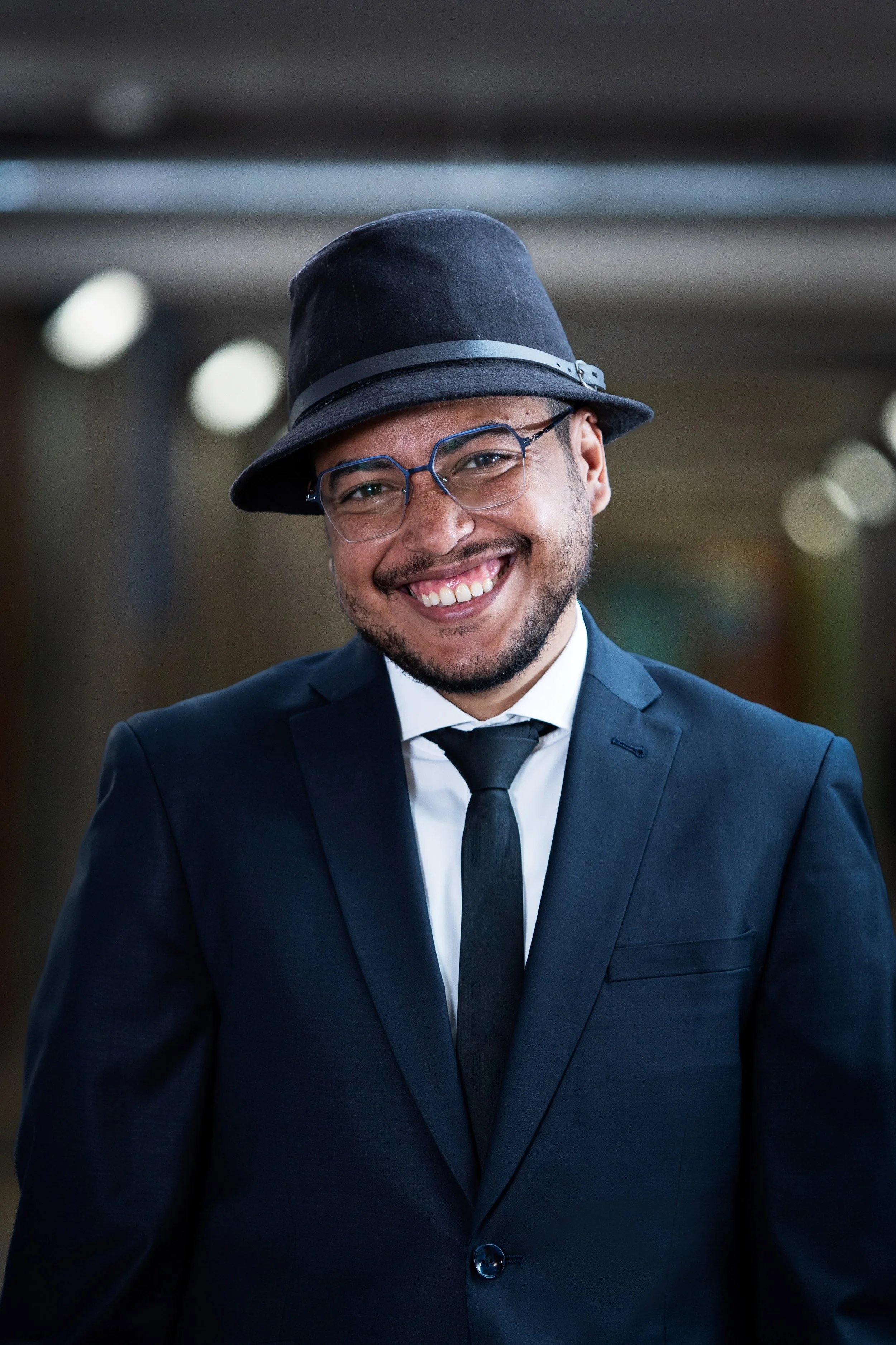 Homme souriant portant un costume noir, une chemise blanche et une cravate noire, avec des lunettes et un chapeau noir, dans un environnement intérieur.