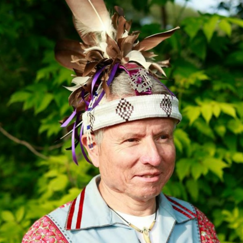 Un homme portant un un costume traditionel autochtone avec des plumes et de rubans, sur un fond de feuillage vert dense.