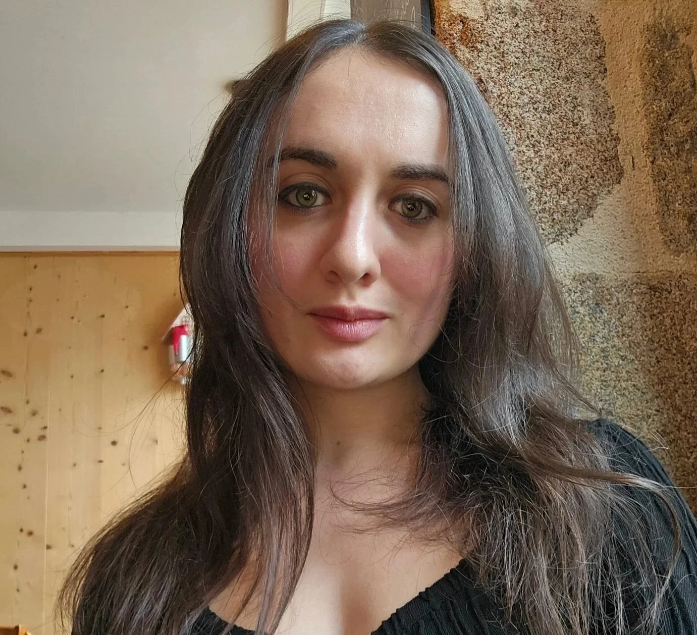 Un portrait d'une femme aux longs cheveux bruns, souriant légèrement, portant un haut noir, avec un mur en pierre en arrière-plan.