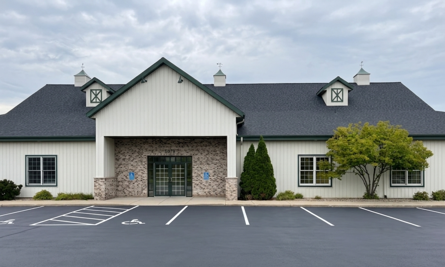 A modern building with a black shingle roof, white siding, and a brick entrance. The building has four small cupolas with weather vanes and large windows. In front, there is a parking lot with several marked handicapped accessible parking spaces, and landscaping with small shrubs and a large tree.