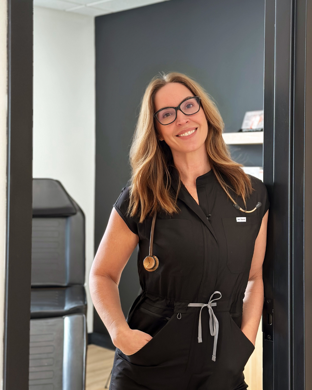 A smiling female healthcare professional wearing glasses, a black medical uniform with a stethoscope around her neck, standing in a clinic or hospital setting.