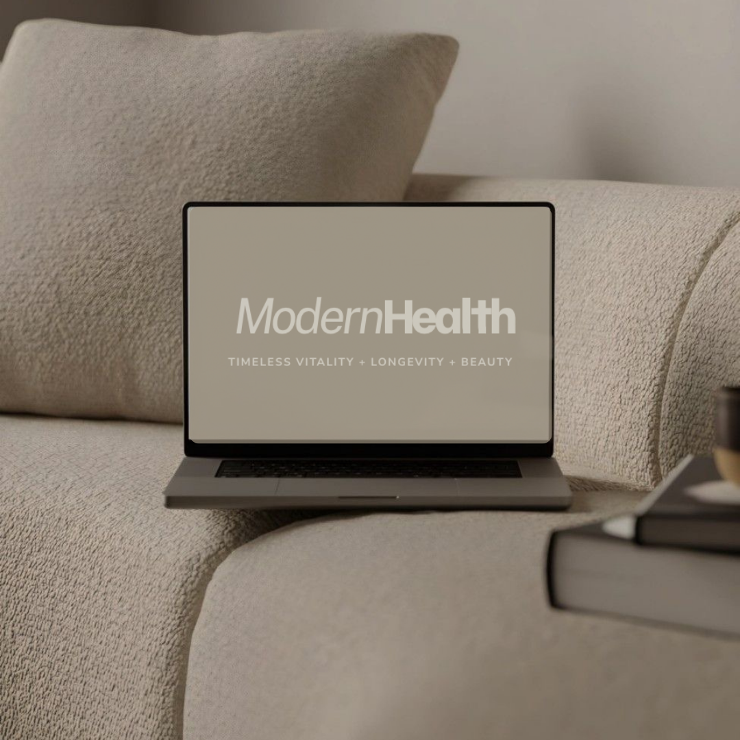 A laptop on a beige sofa displays the logo of ModernHealth with the tagline 'Timeless Vitality + Longevity + Beauty'.