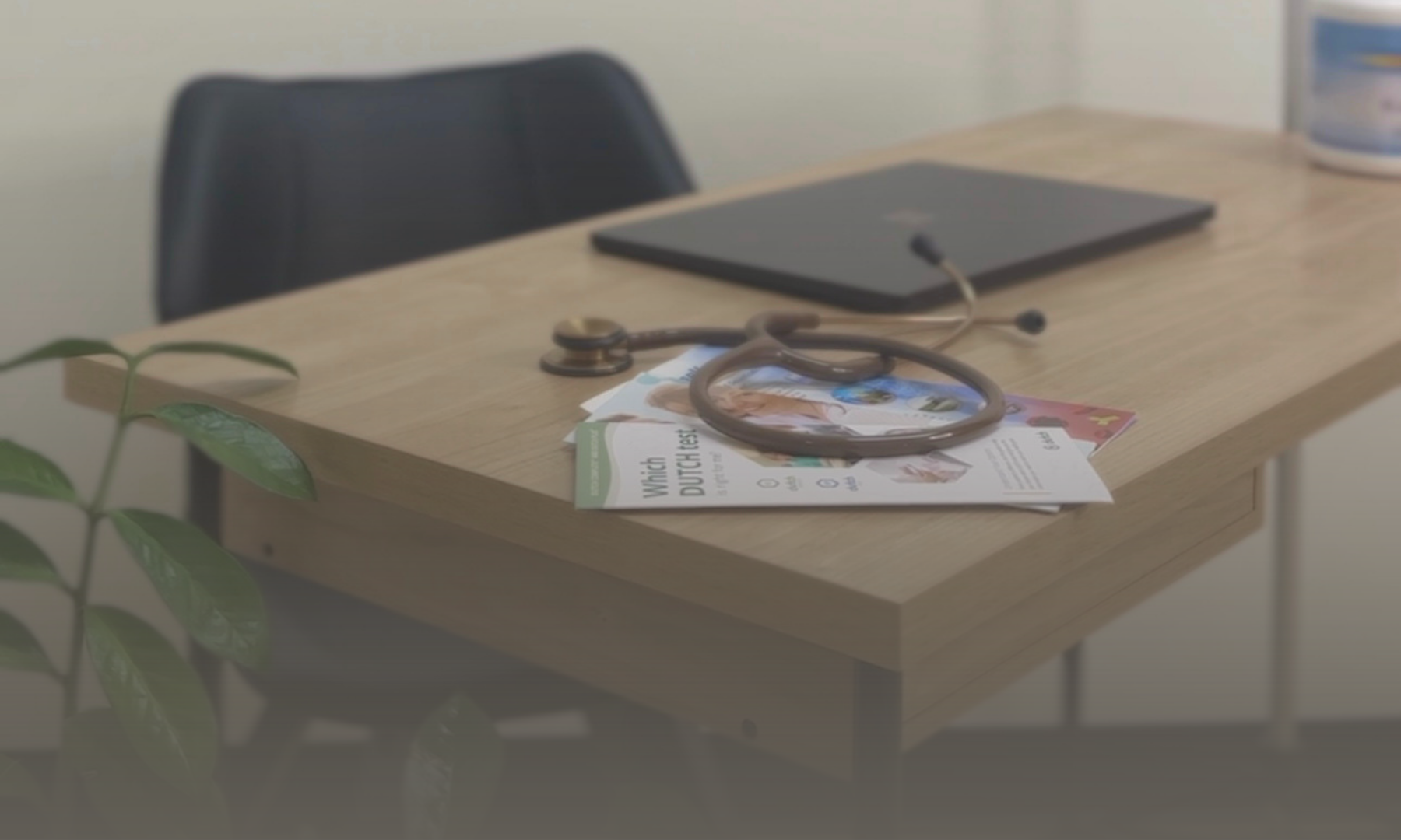 A wooden desk with a stethoscope, a gray laptop, medical pamphlets, and a partially visible potted plant in the foreground, with a blue and white container on the right side of the desk.