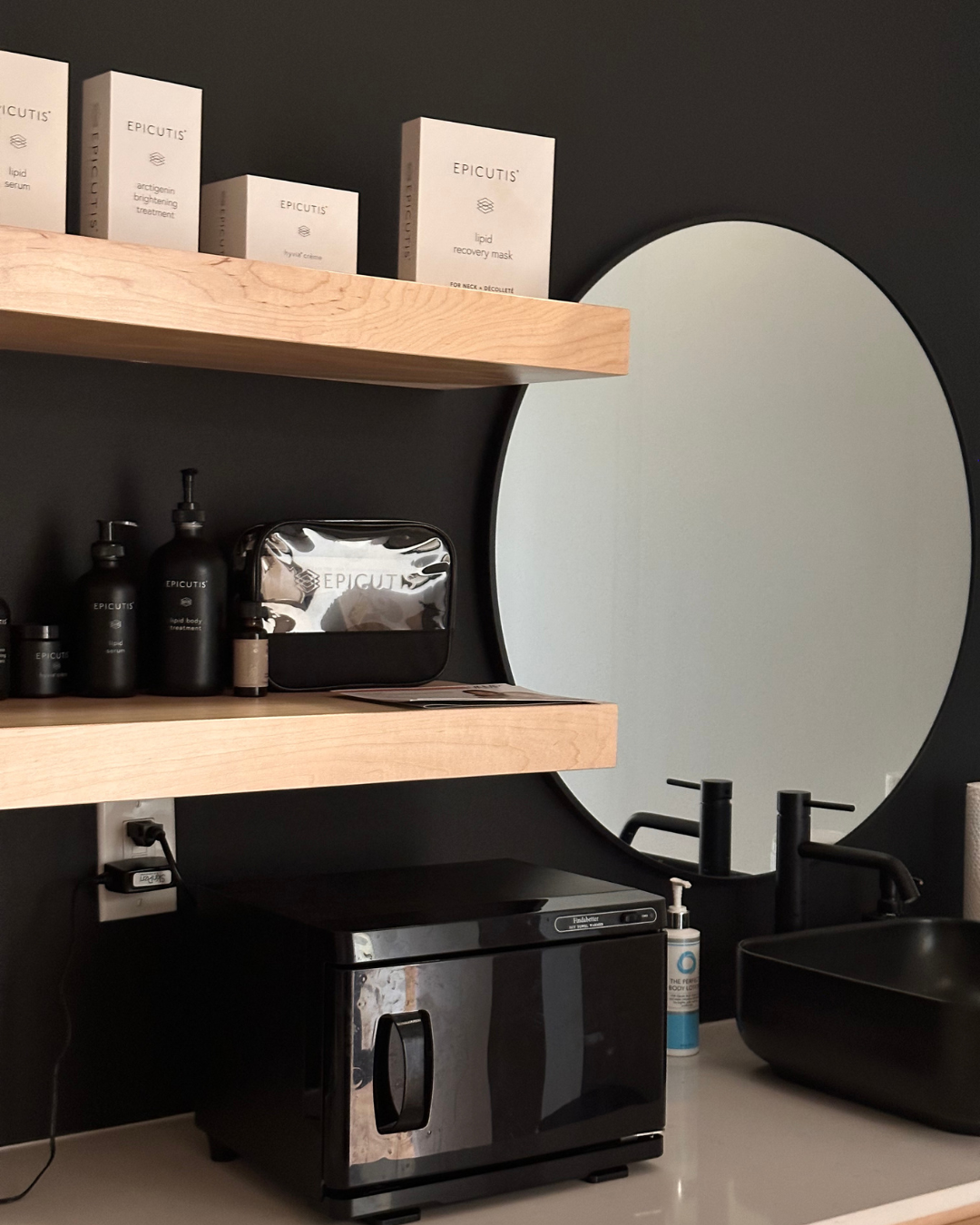 Bathroom with black walls, a round mirror, black sink, black soap dispensers, a black mini refrigerator, and wooden shelves holding skincare products and a silver toiletry bag.