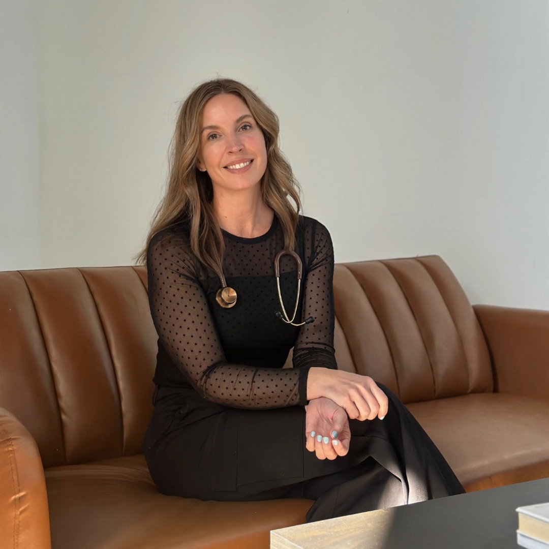 A woman with long wavy brown hair, wearing a black dotted sheer long-sleeve top and black pants, sitting on a brown leather couch with a stethoscope around her neck, smiling at the camera.