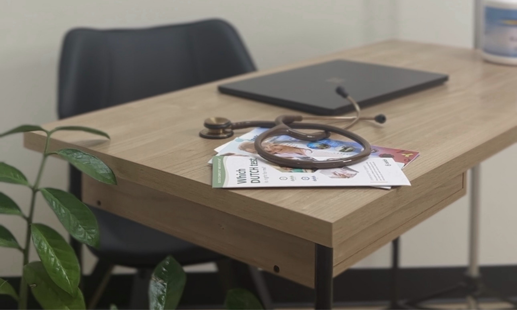 Medical stethoscope resting on papers and pamphlets on a wooden table, with a laptop and a black chair in the background, and a green plant in the foreground.