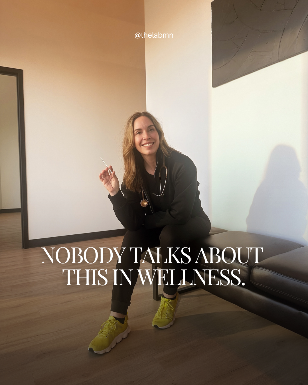 A woman sitting on a black leather bench in a wellness clinic, smiling with a stethoscope around her neck, holding a syringe, wearing black athletic pants, yellow sneakers, and a black hoodie. The wall behind her is white and part of an abstract black art piece. Text overlay reads, "NOBODY TALKS ABOUT THIS IN WELLNESS."