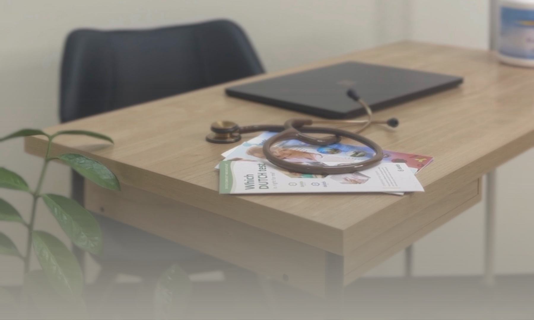 A wooden desk with a black chair behind it. On the desk, there are scattered papers, a stethoscope, a closed laptop, and a container. A green plant is partially visible in the foreground.