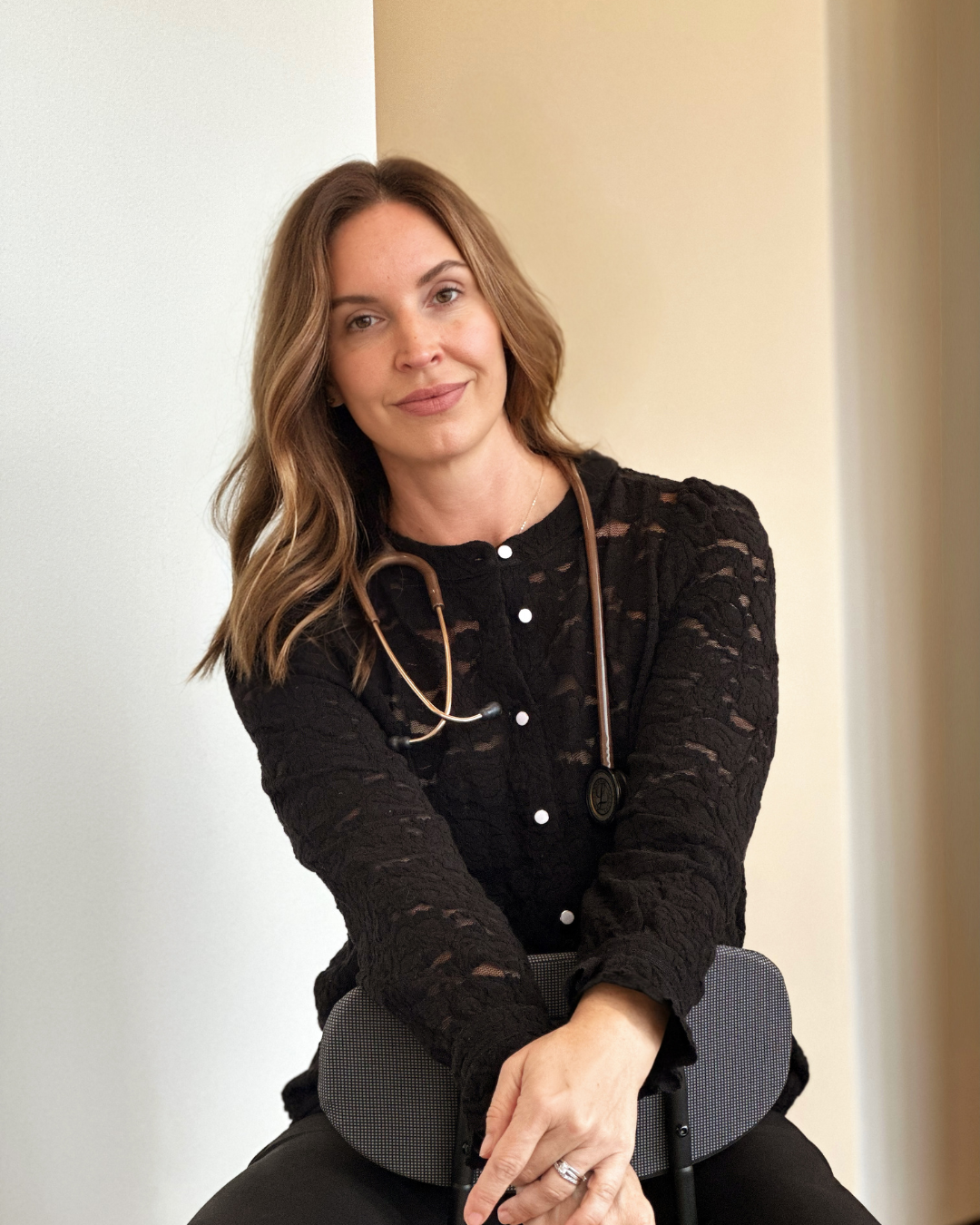 A woman with light skin, medium-length wavy brown hair, and a slight smile, sitting with her arm resting on her knee. She is wearing a black lace long-sleeve top with white buttons, a necklace, and a stethoscope around her neck. She appears to be in a professional setting.