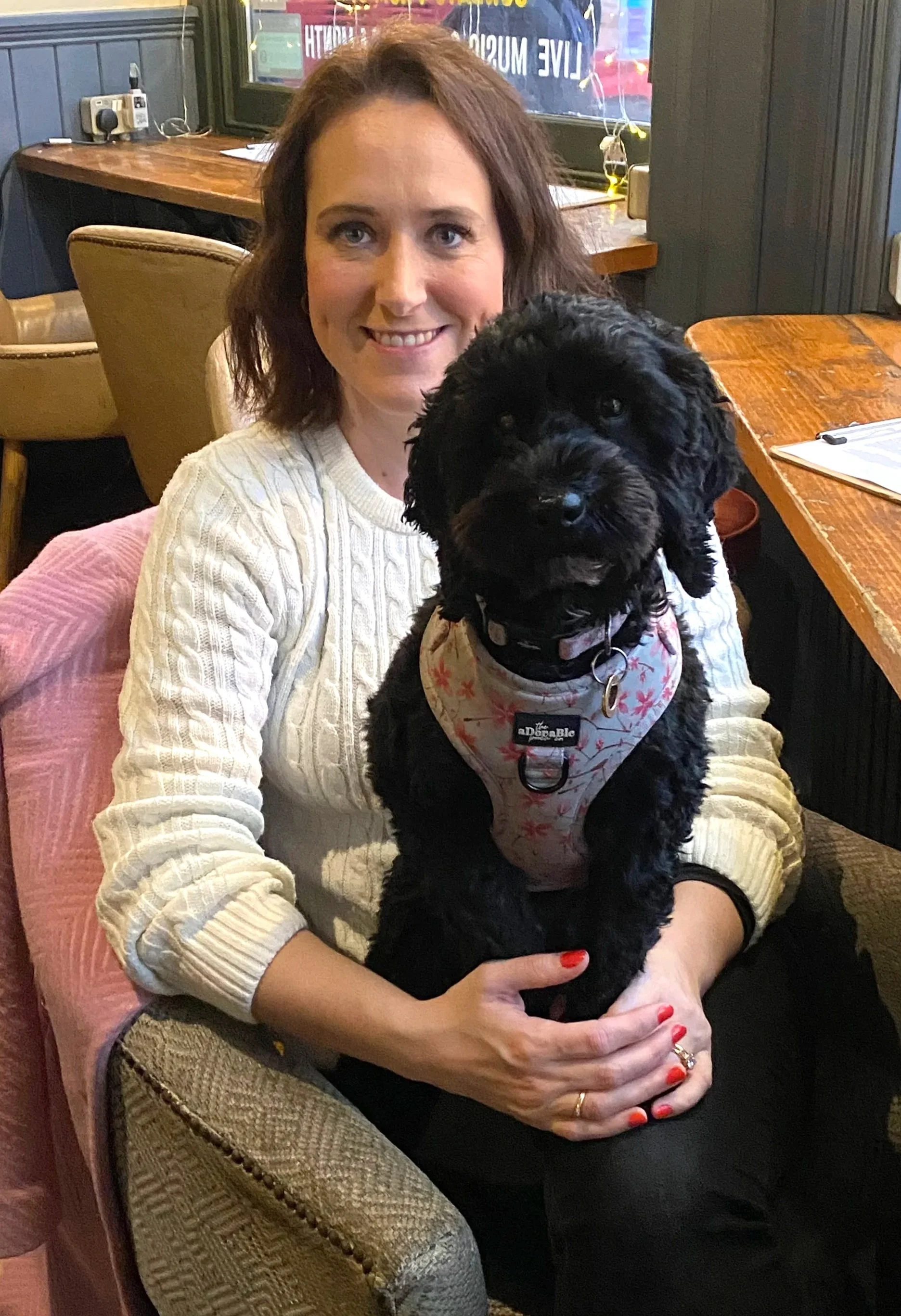 A woman with brown hair and a beige cable-knit sweater holding a black dog with curly fur and a floral bandana indoor at a cozy restaurant or cafe.