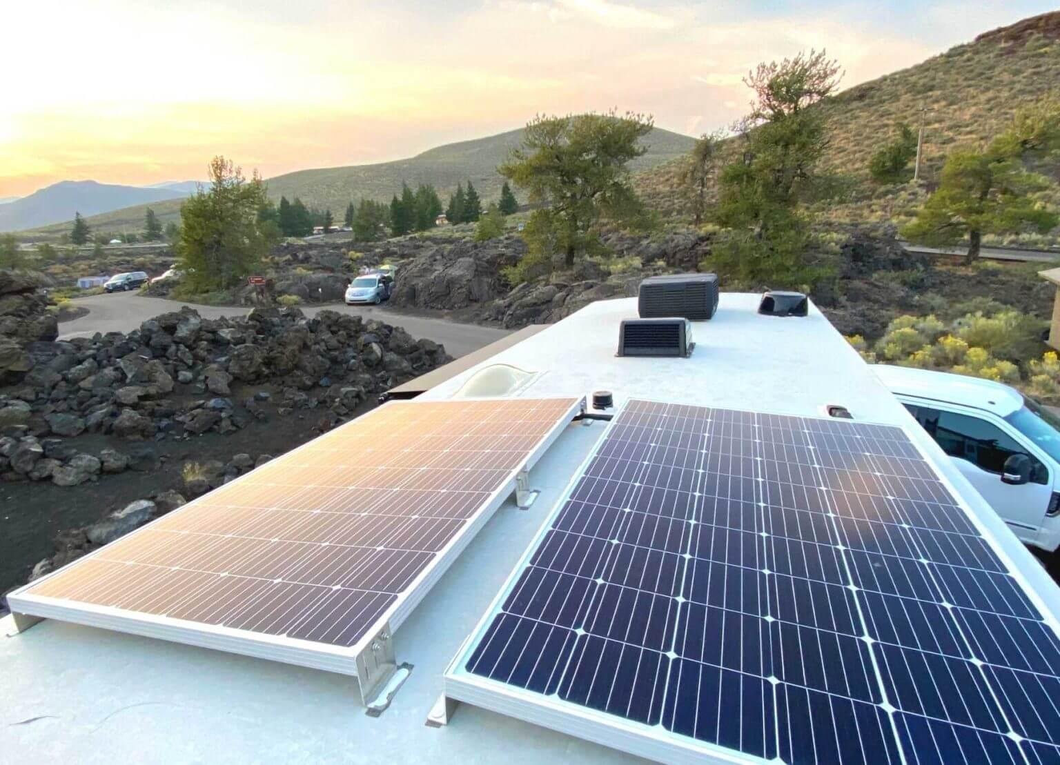 Solar panels installed on the roof of a vehicle in a mountainous area at sunset, with multiple parked cars and trees in the background.