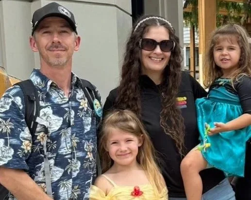Four people standing together outdoors, including a man wearing a Hawaiian shirt and a backpack, a woman with long curly hair in sunglasses, and two young girls, one in a yellow dress and the other in a blue dress.