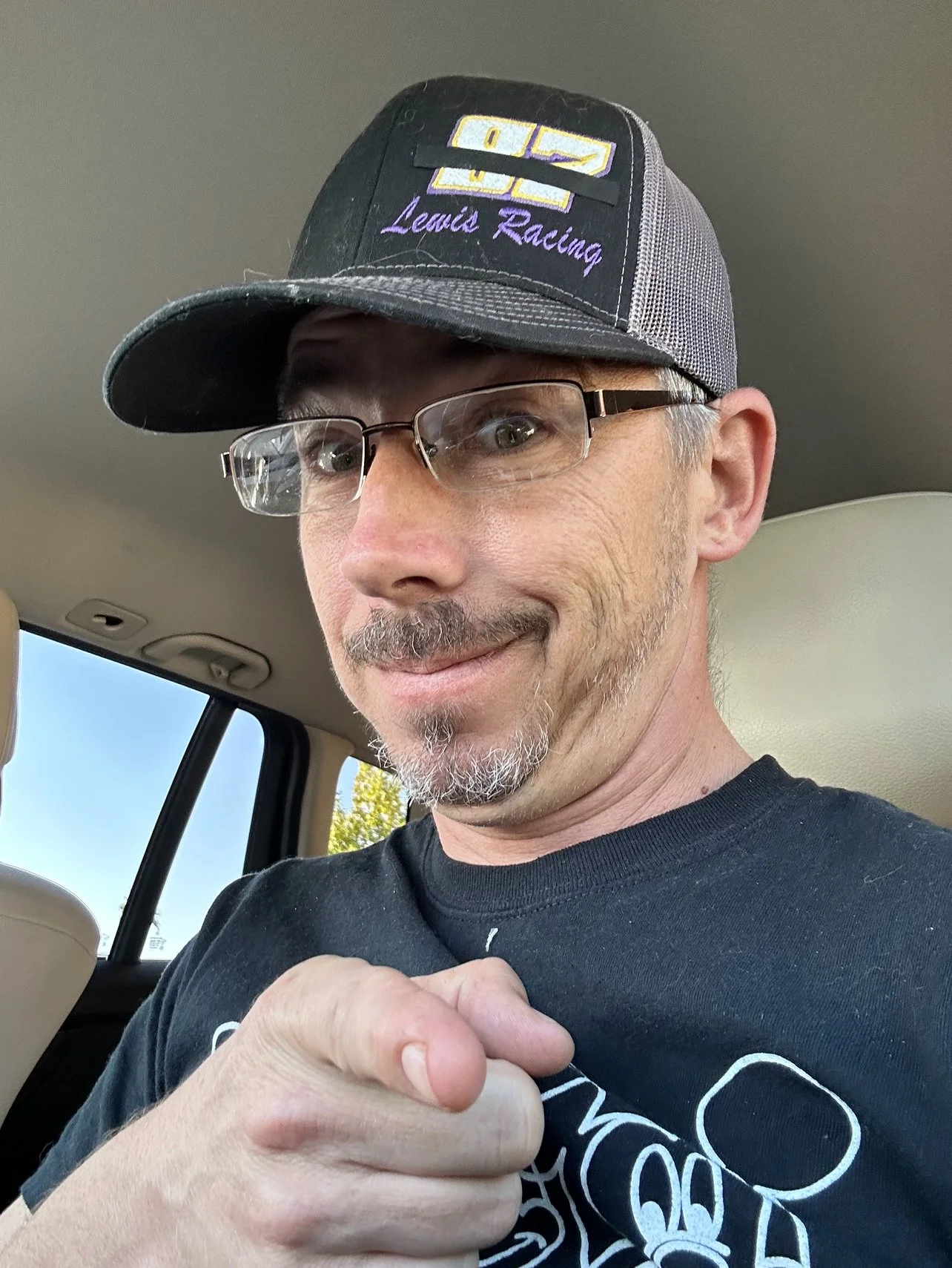 A man with glasses and a gray goatee wearing a black t-shirt and a black baseball cap with purple and yellow embroidery that says "Lewis Racing". He is smiling and pointing with his hand.