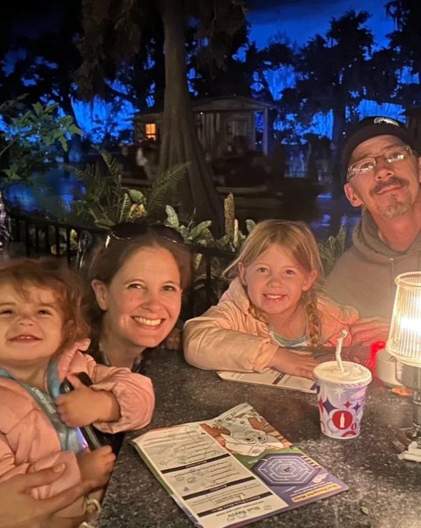 Family enjoying dinner at an outdoor restaurant at night, with two children and two adults smiling at the camera.