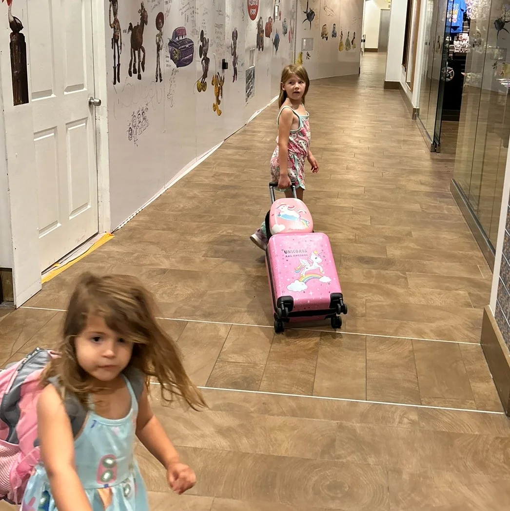 Two young girls in a hallway, one pulling a pink suitcase with unicorn and rainbow designs, the other carrying a backpack.