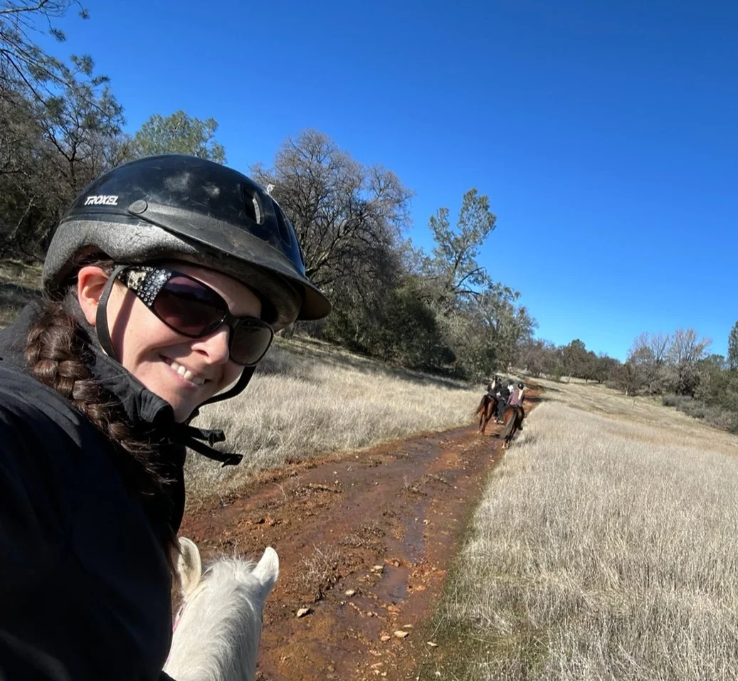 Person wearing a helmet and sunglasses riding a horse on a dirt trail in a field, with two more people riding horses in the background.