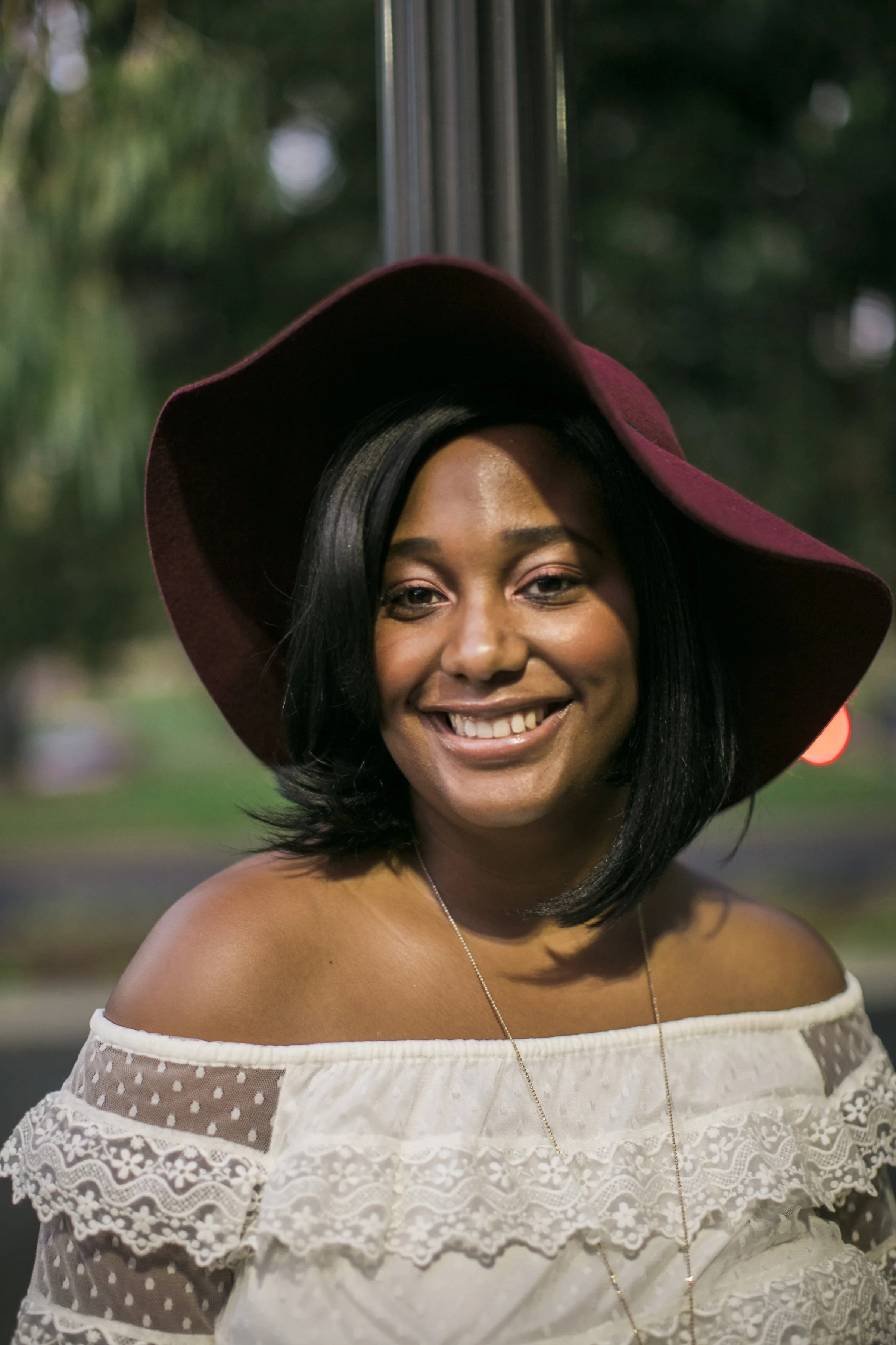 A smiling woman with black hair wearing a burgundy wide-brim hat and an off-shoulder white lace top.