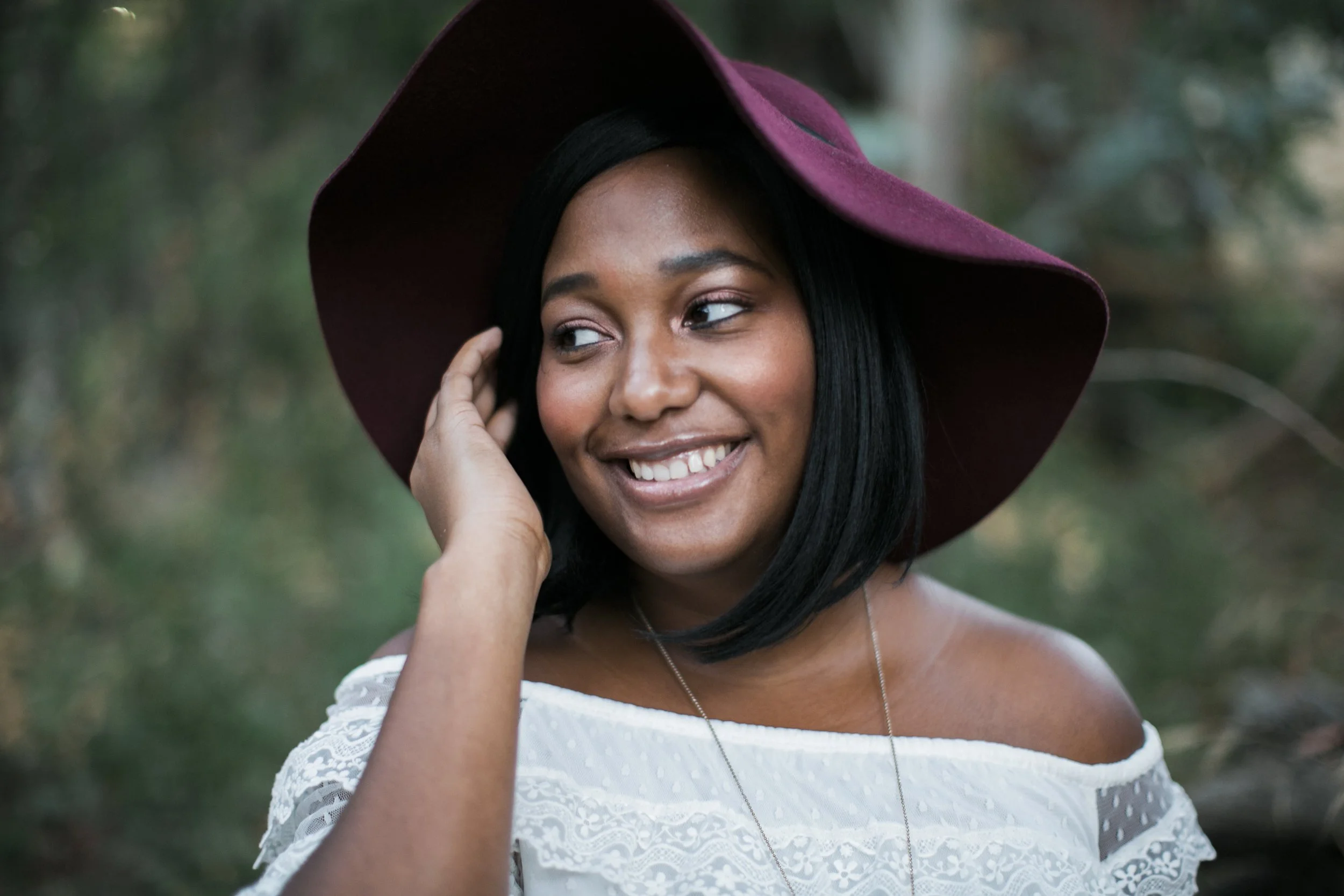 A woman wearing a maroon wide-brimmed hat and a white lace off-the-shoulder top, smiling and looking to the side, outdoors with blurred greenery in the background.