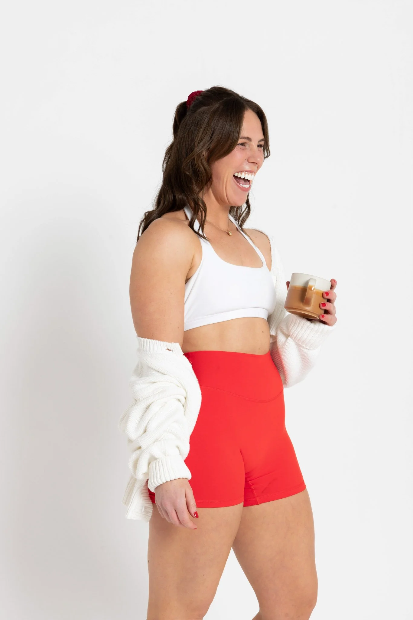 A woman with long brown hair, wearing a white sports bra and red shorts, holding a beige ceramic mug, laughing, with a white cardigan draped over her shoulders, standing against a plain white background.