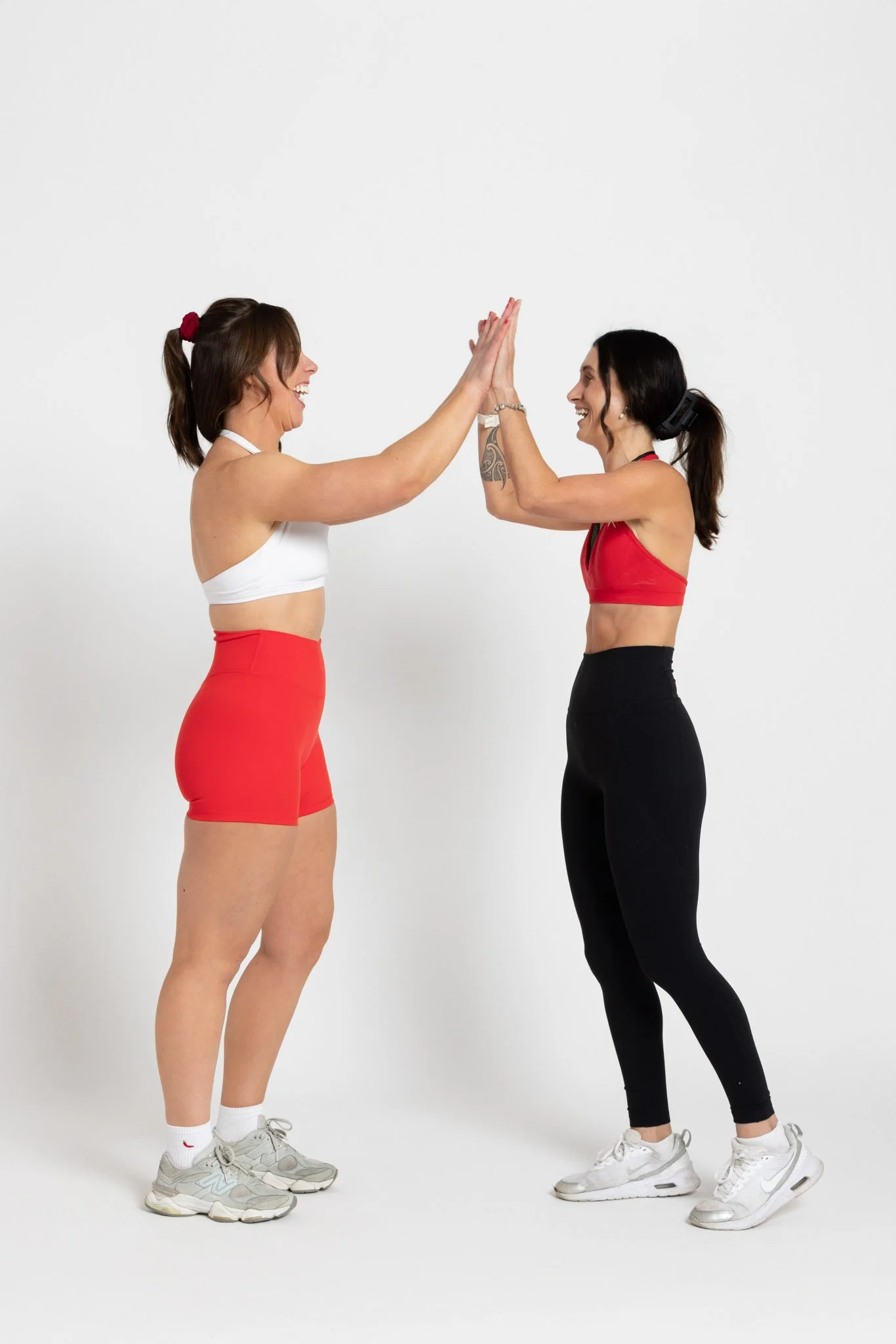 Two women in athletic outfits, smiling and giving high five in a white studio. One woman is wearing a white sports bra and red shorts, white socks, and gray sneakers. The other woman is wearing a red sports bra, black leggings, and white sneakers, with a black headband and tattoo on her left arm.