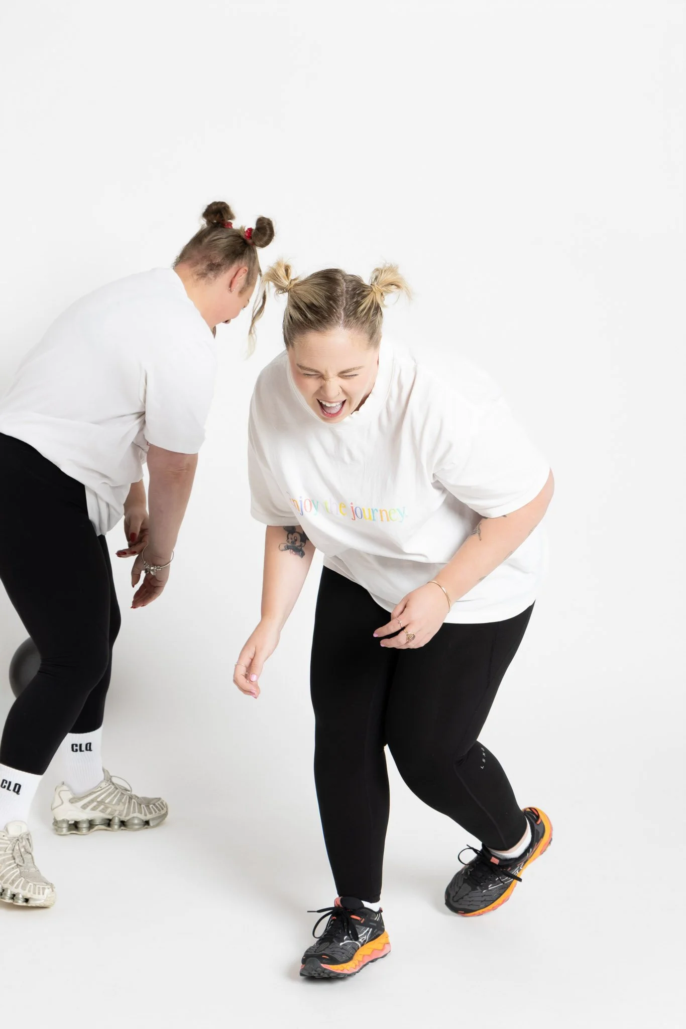 Two women with blonde hair styled in buns are placing their feet together and leaning towards each other, appearing playful and engaged with each other.