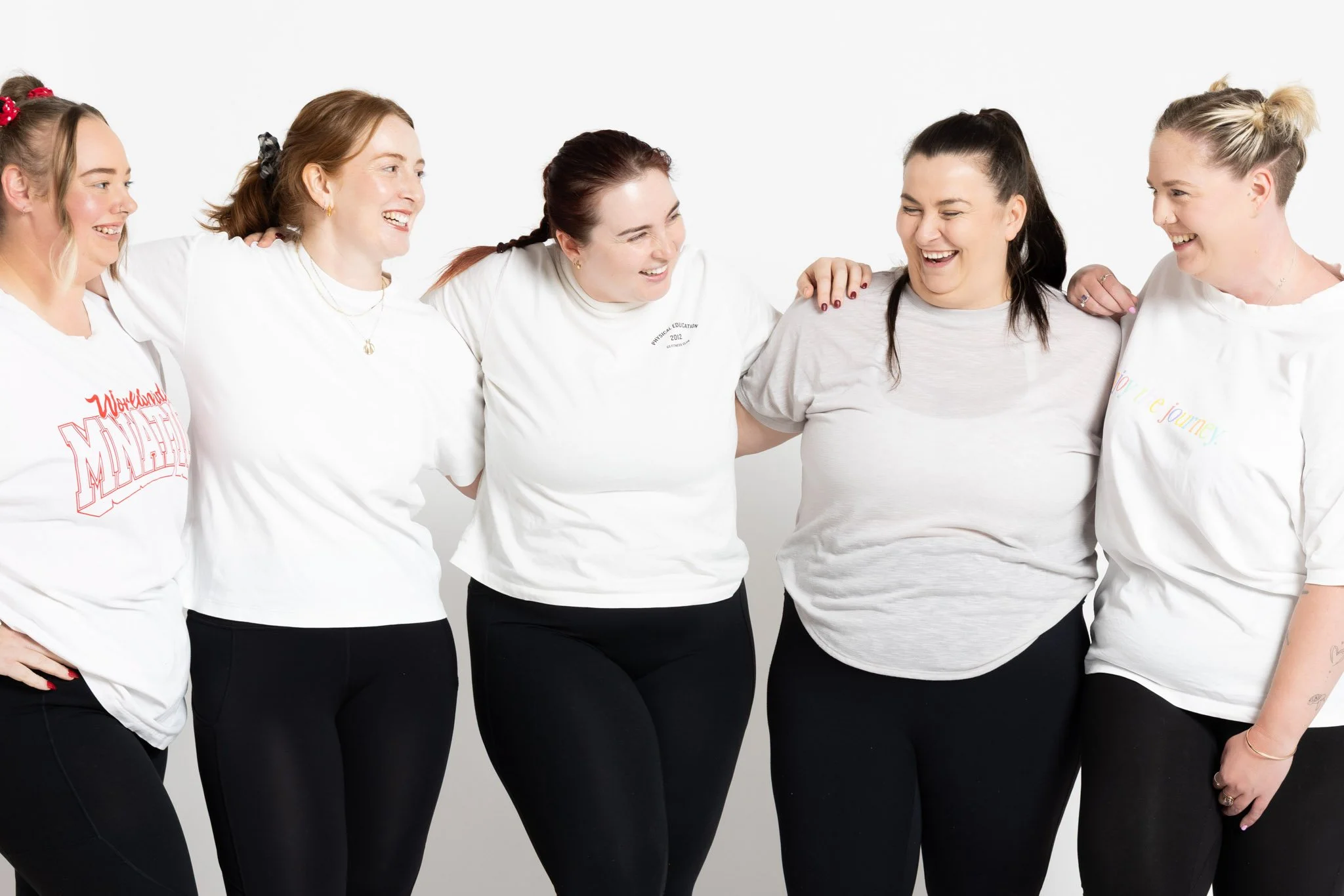 Six women standing close together with arms around each other, smiling and laughing, against a plain white background.
