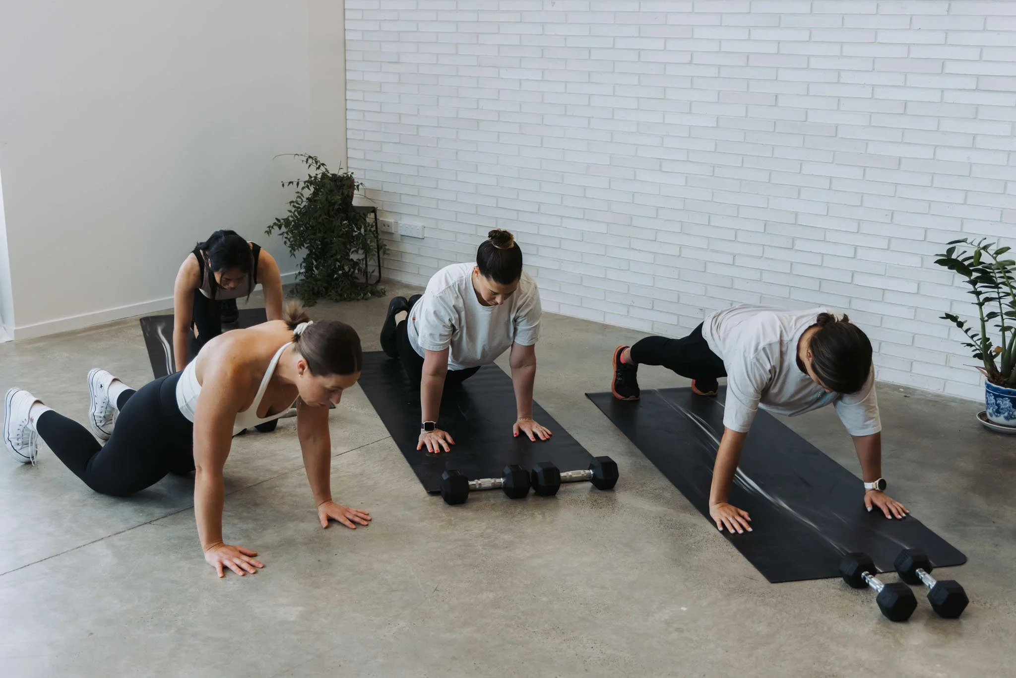Four women doing a workout in a gym with black mats, black dumbbells, and a white brick wall, while a woman in a beige t-shirt leads the exercise.