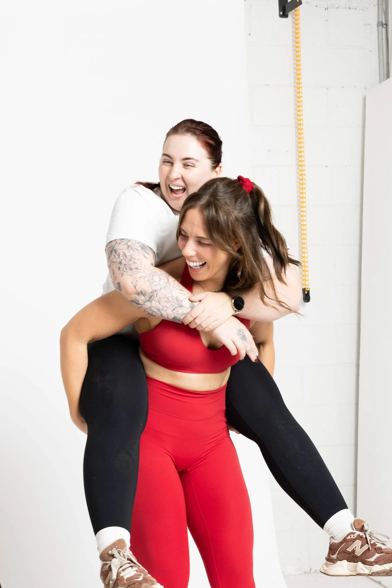 Two women engaging in a playful piggyback ride indoors. The woman in the red workout outfit is carrying the woman in black leggings, who is smiling and wearing a white shirt. They appear happy and are in a gym or fitness studio with a white wall and a measuring tape on the wall.
