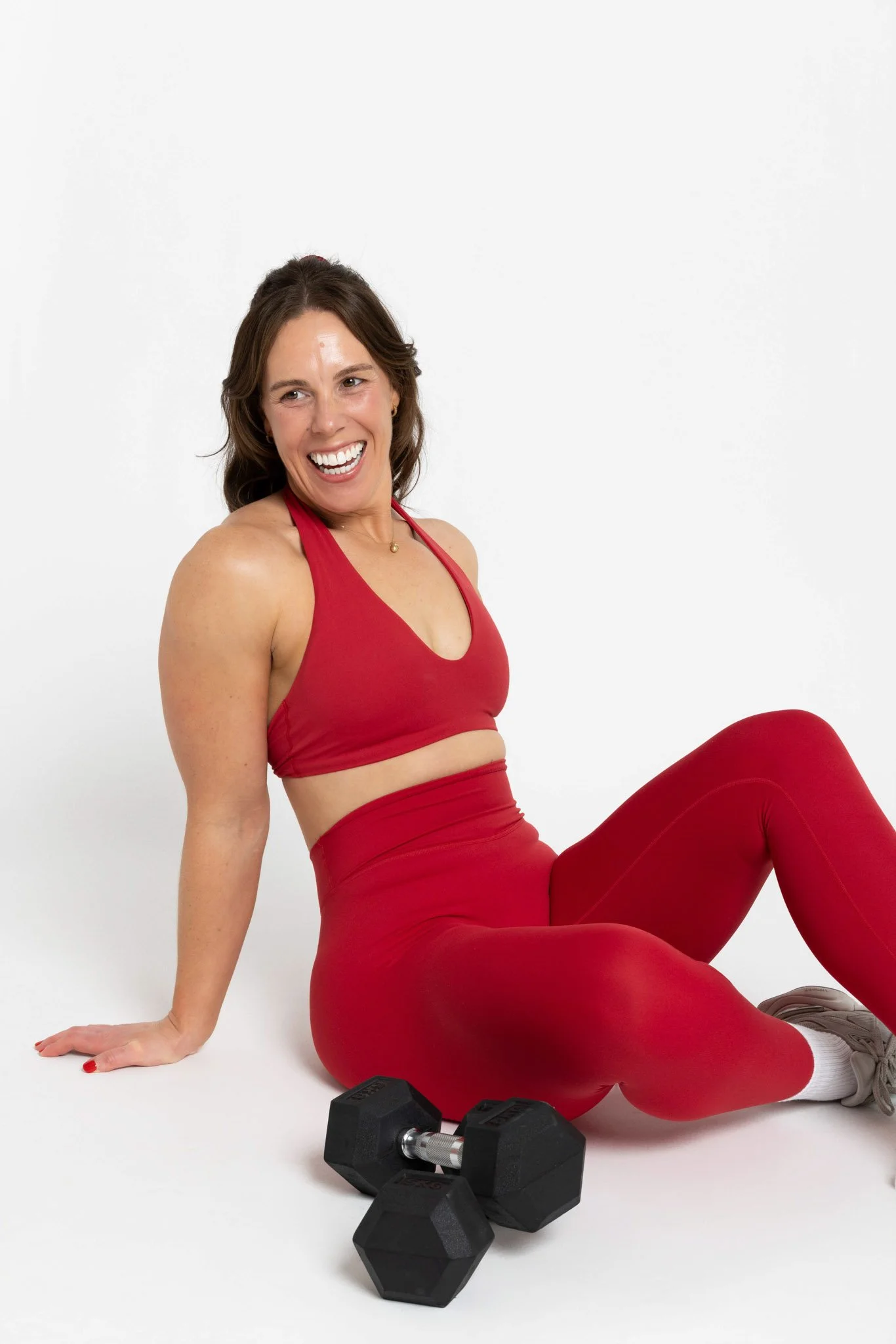 Woman in red workout clothing sitting on the floor, smiling, with dumbbells and a water bottle nearby.