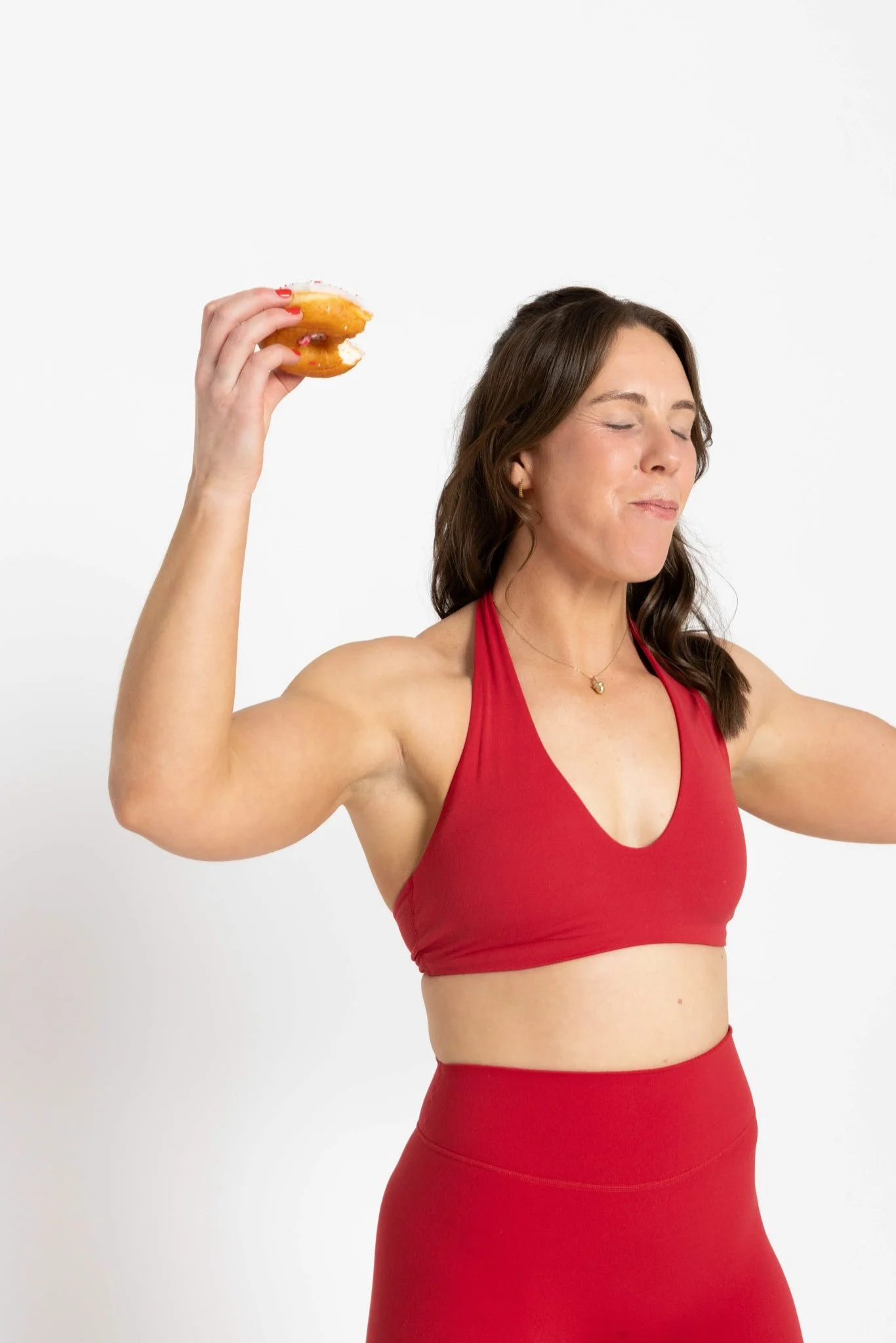 A woman in a red sports bra and matching athletic high-waisted leggings, with long brown hair, is holding a bitten glazed donut with pink sprinkles in her right hand while closing her eyes and smiling.