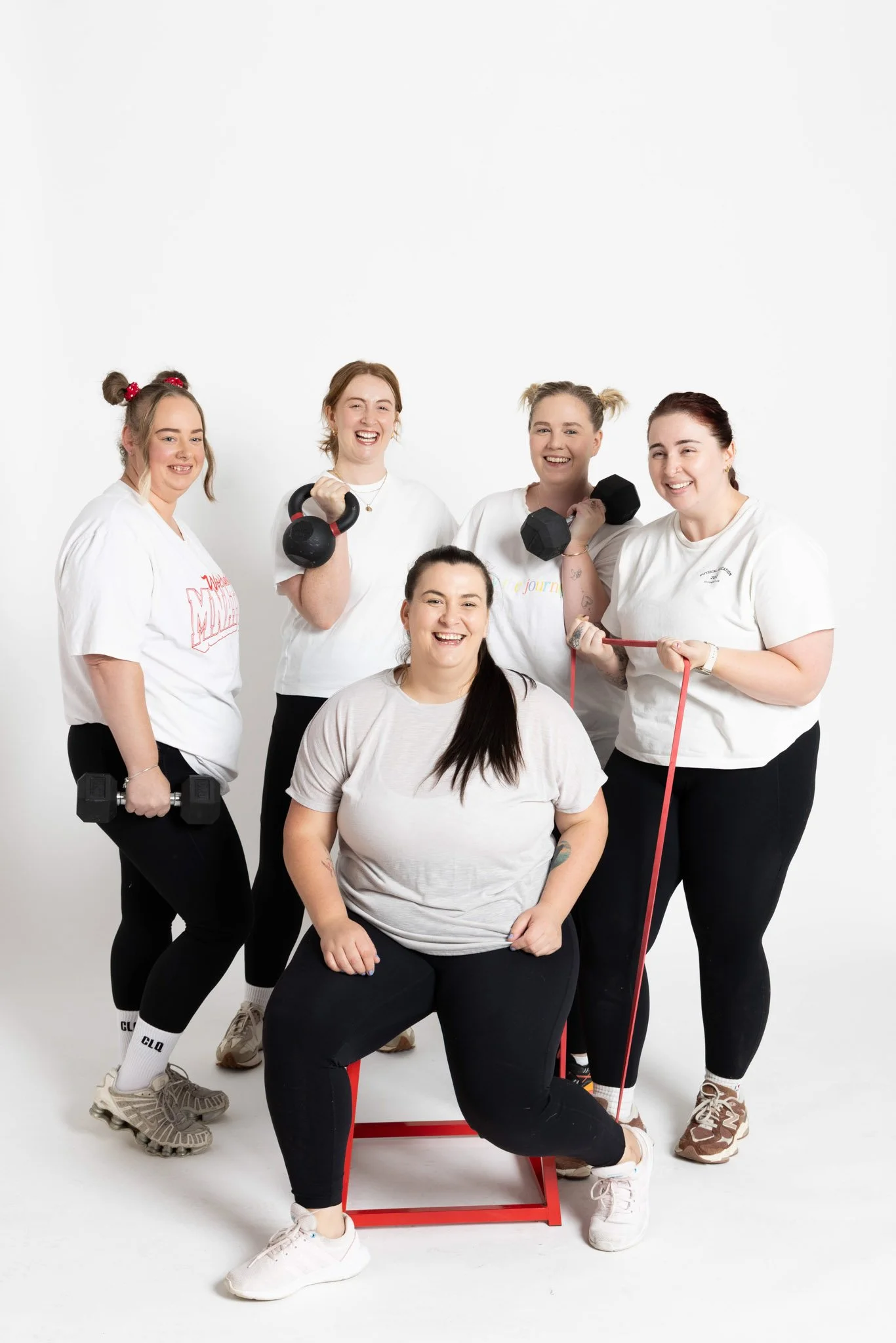 Group of five diverse women in athletic wear smiling and holding workout equipment like dumbbells and resistance bands against a plain white background.
