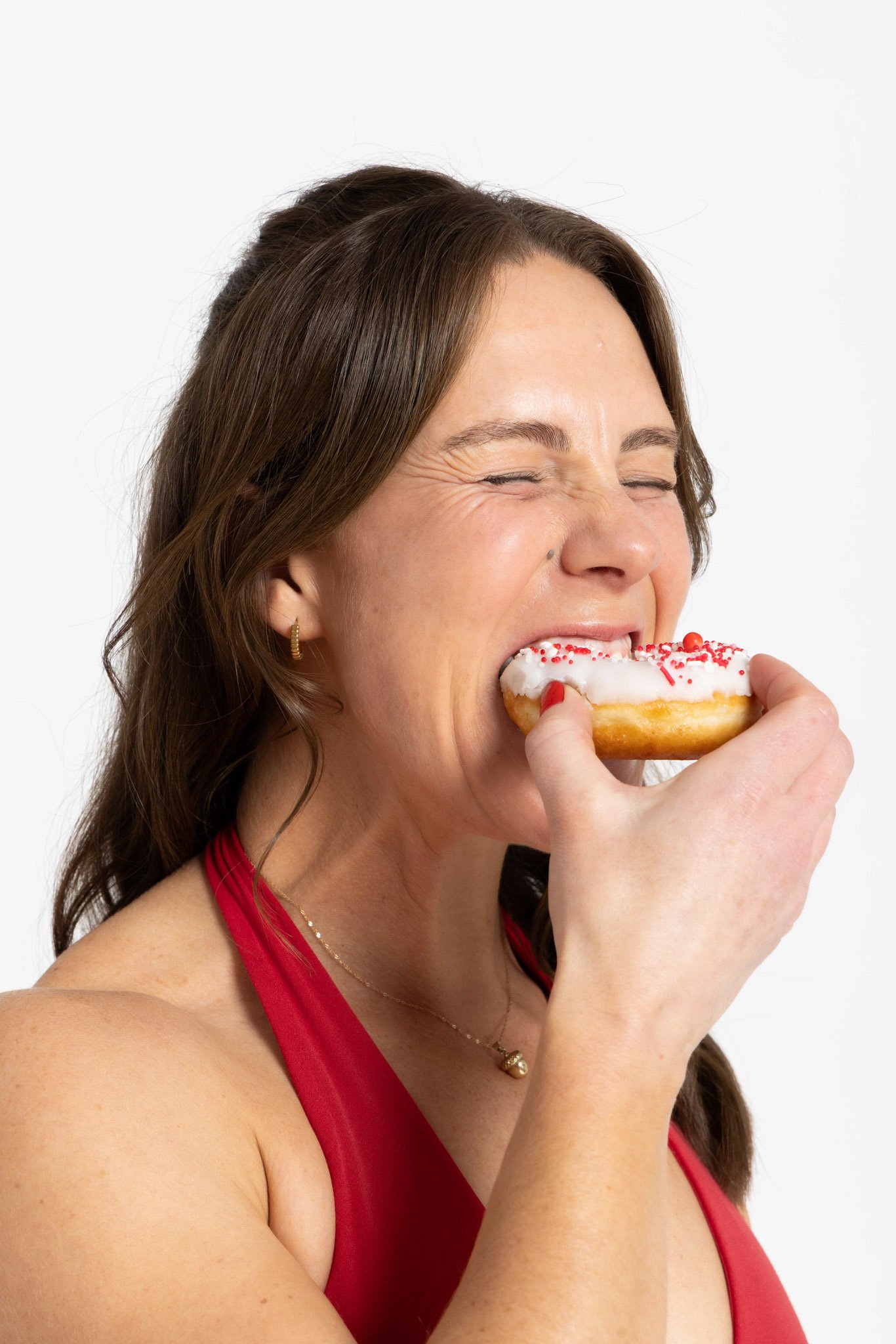 A woman with brown hair, wearing a red dress and gold jewelry, is smiling with her eyes closed while holding and about to bite a decorated donut.