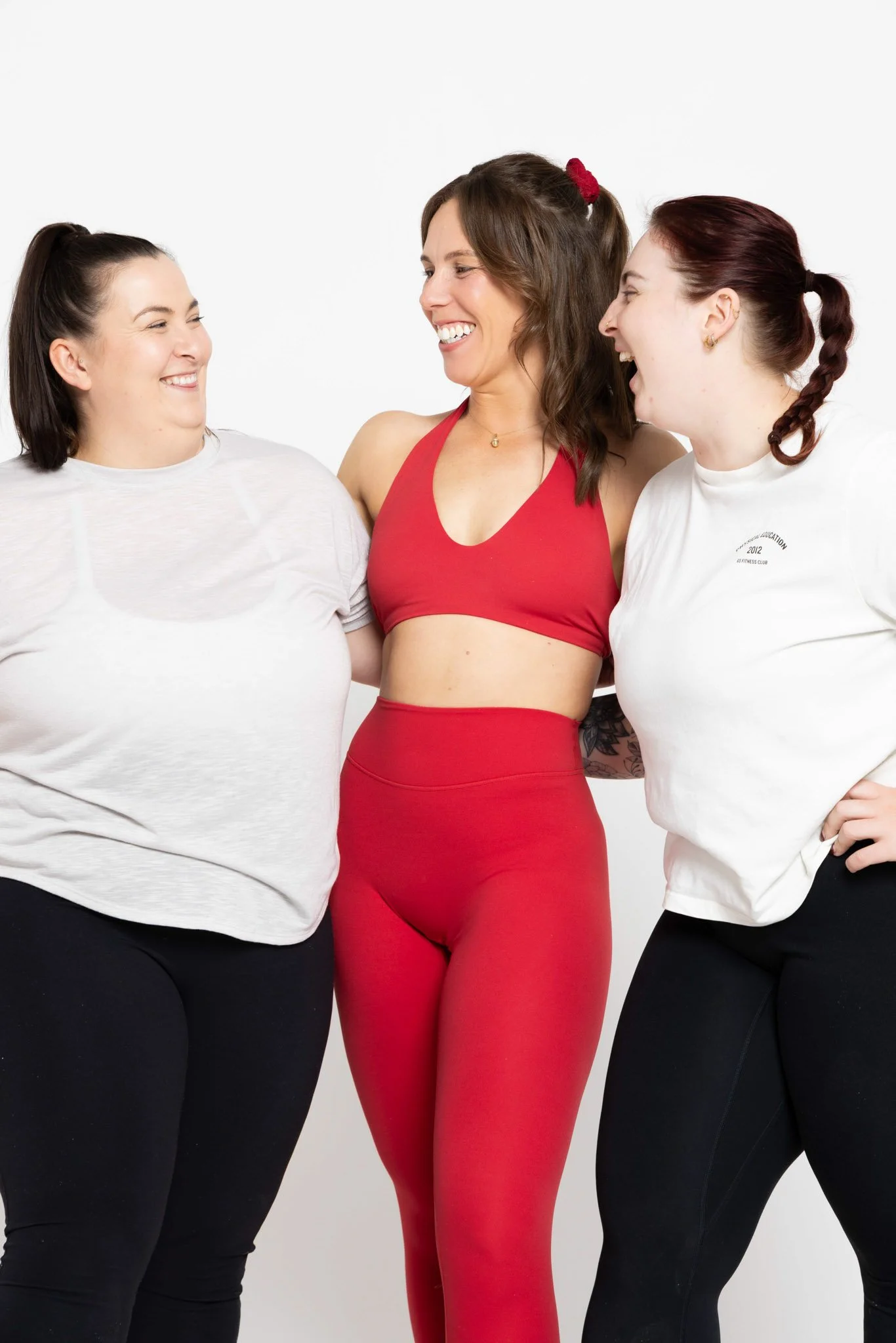 Three women smiling and looking at each other, standing close together against a plain white background, one in red workout clothing and two in white t-shirts and black pants.