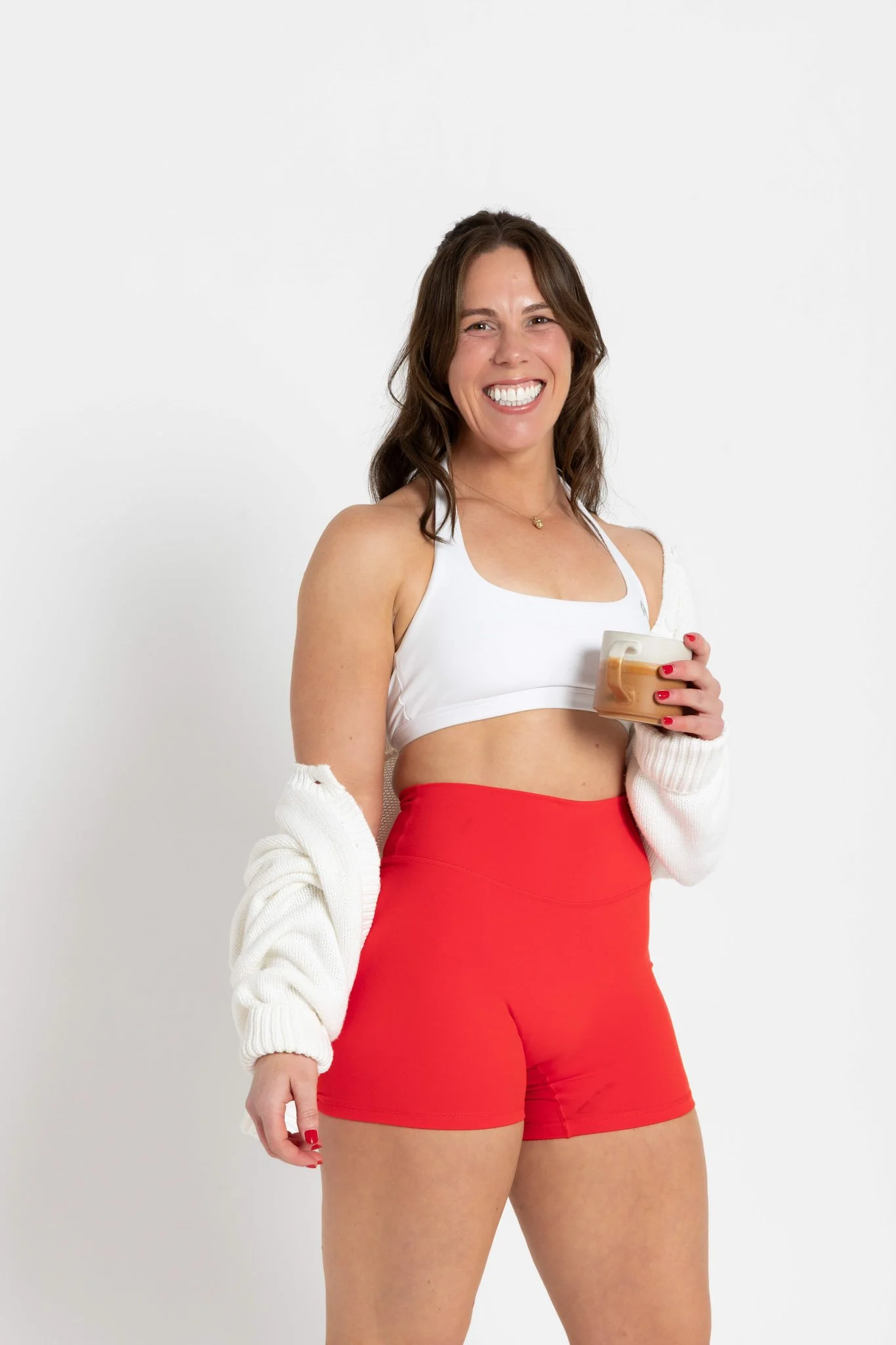 A woman with brown hair, smiling while holding a glass of coffee, wearing a white sports bra, bright red shorts, and a white cardigan.