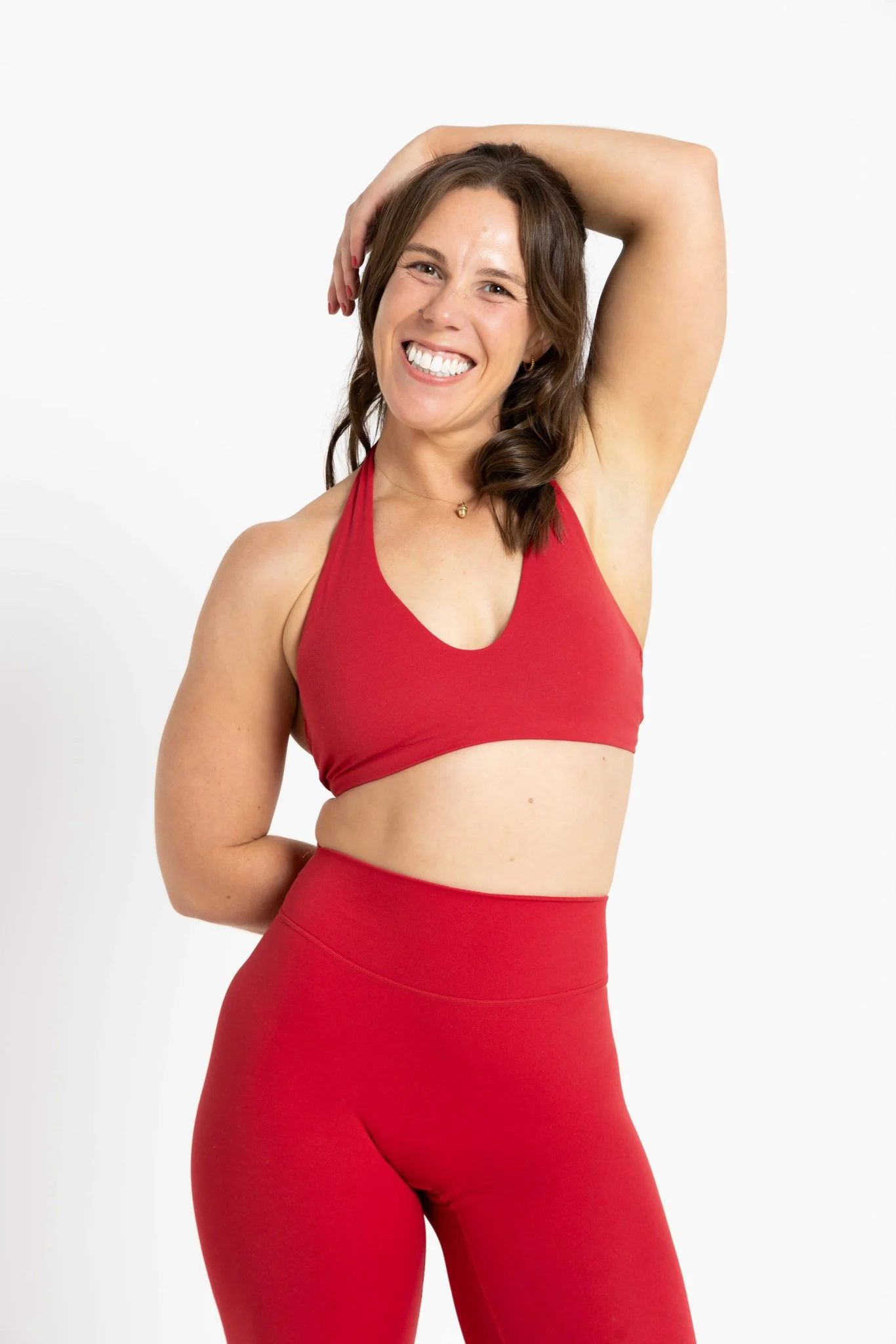 A woman with brown hair smiling, wearing a red sports bra and matching high-waisted leggings, posing with one arm bent and hand resting on her head against a plain white background.