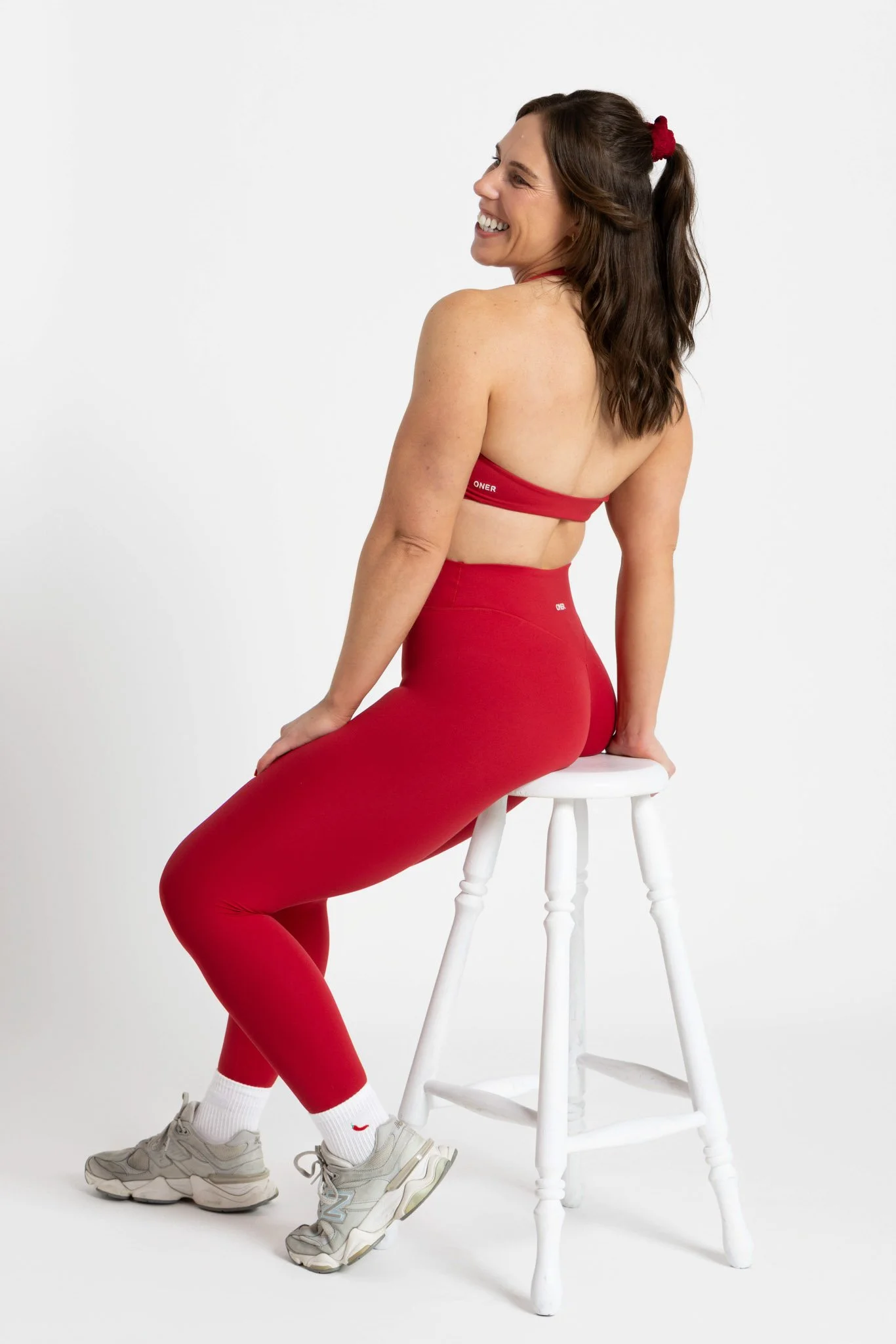 A woman with long brown hair tied with a red scrunchie, sitting on a white stool, wearing red workout leggings, a red sports bra, white sneakers, and white socks, smiling and looking over her shoulder against a plain white background.