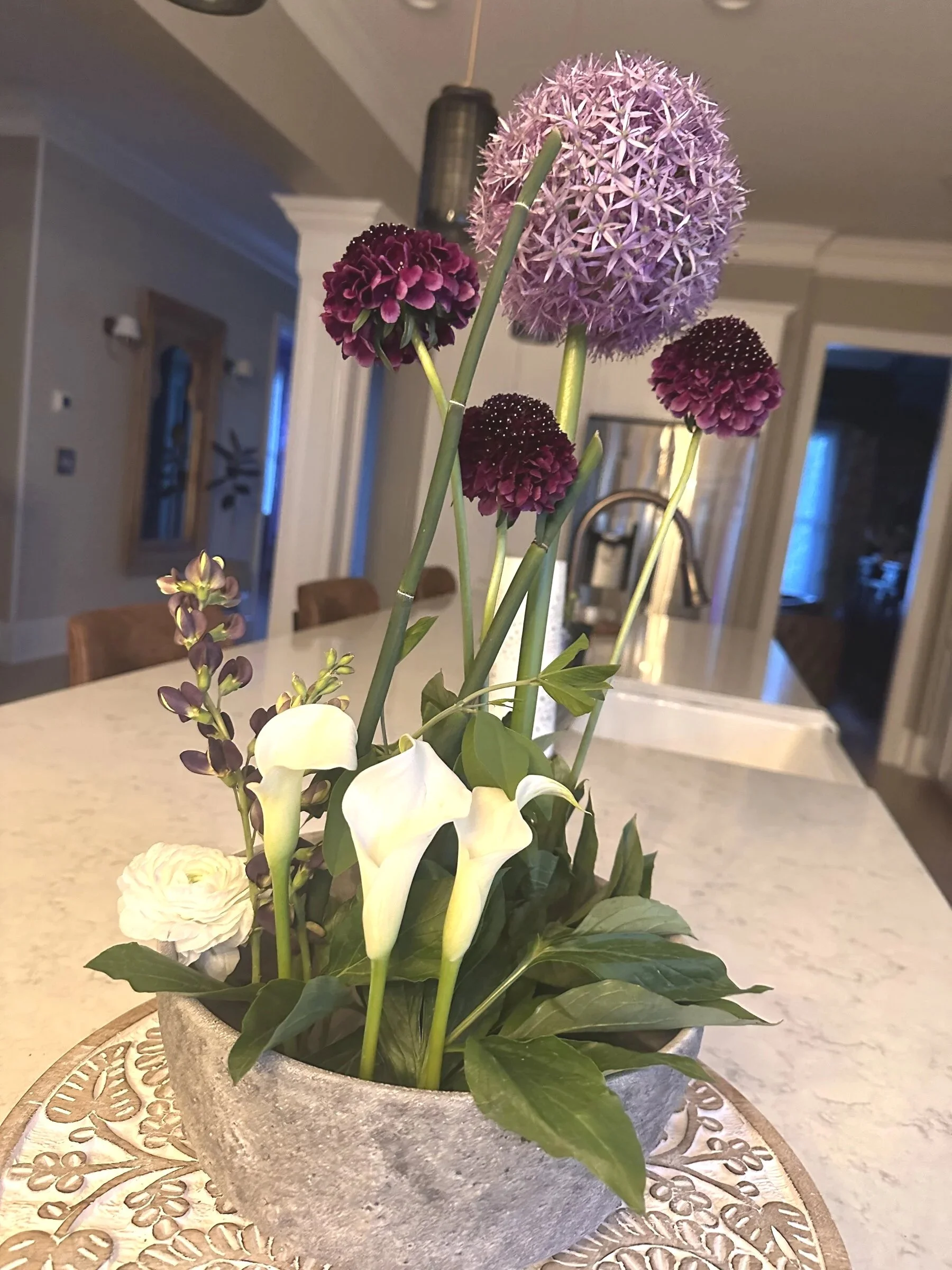 A flower arrangement with allium and white calla lilies in a round, gray, textured pot on a decorative tray in a kitchen.