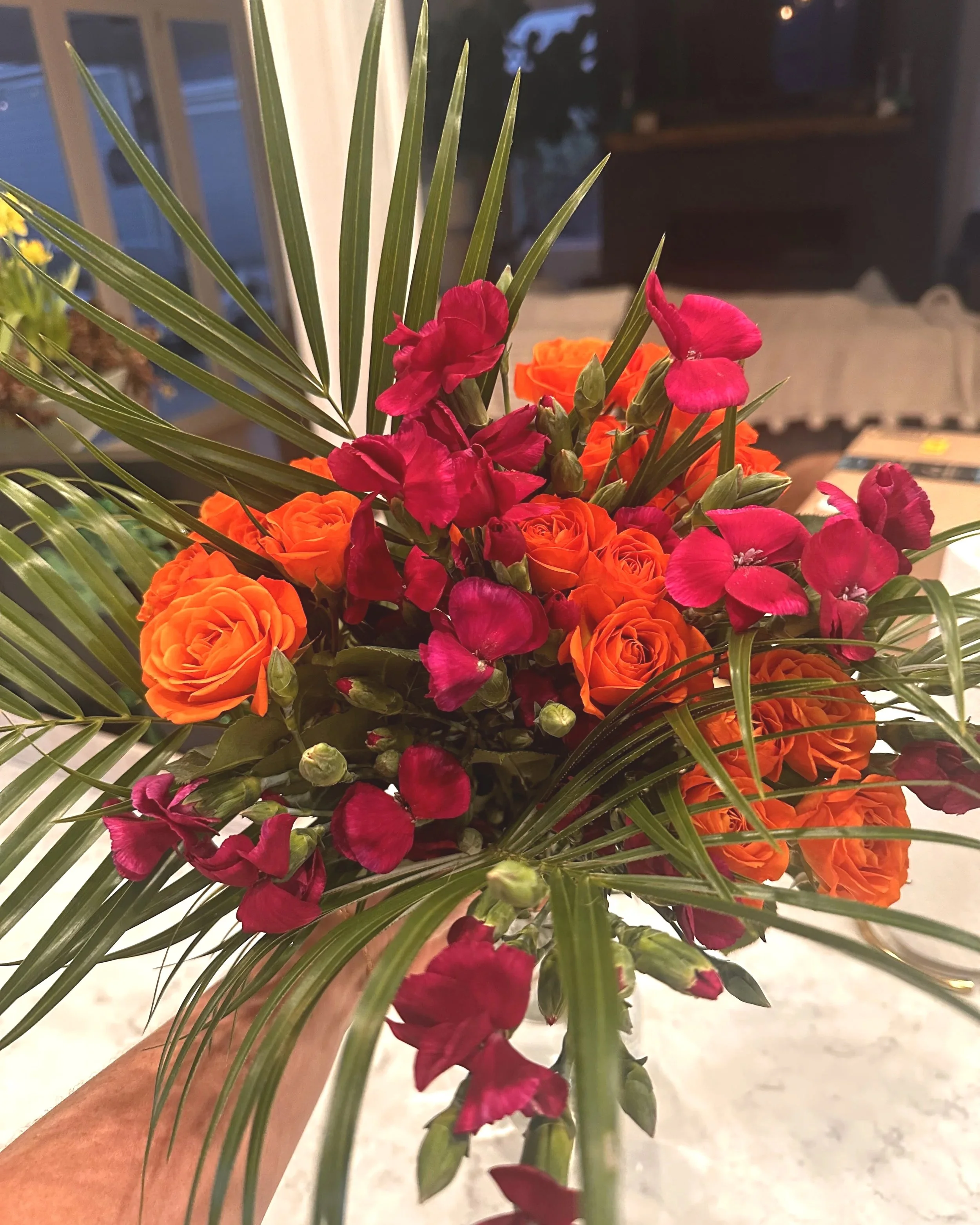 A hand holding a bouquet of mixed flowers, including red and orange roses, fuchsia flowers, and green palm leaves, with a blurred indoor background.