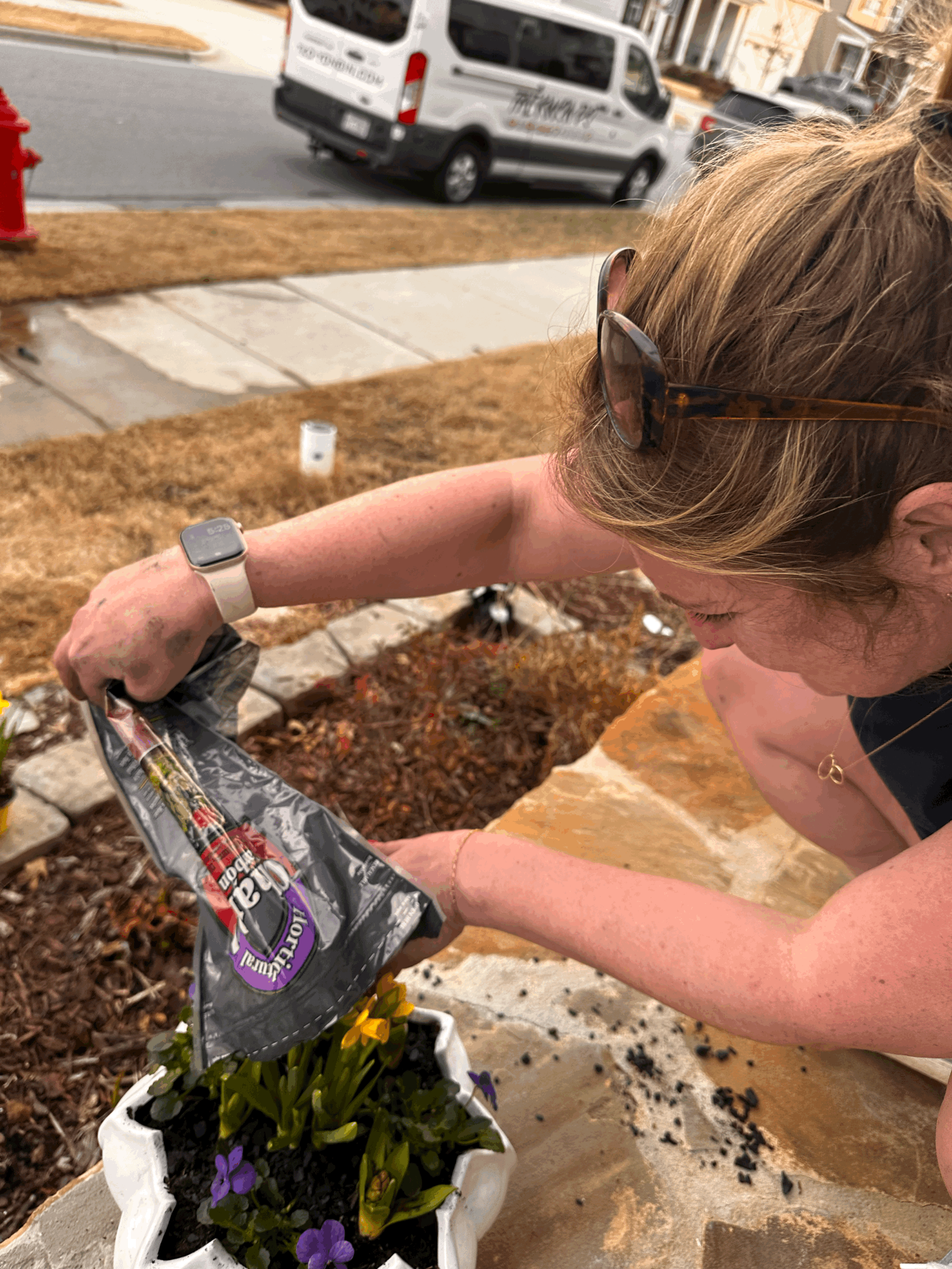 A woman planting purple flowers in a white flower pot on a stone surface outside. She is wearing sunglasses, a watch, and a bracelet, with dirt on her arms. In the background, there is a sidewalk, a parked van, and other vehicles.